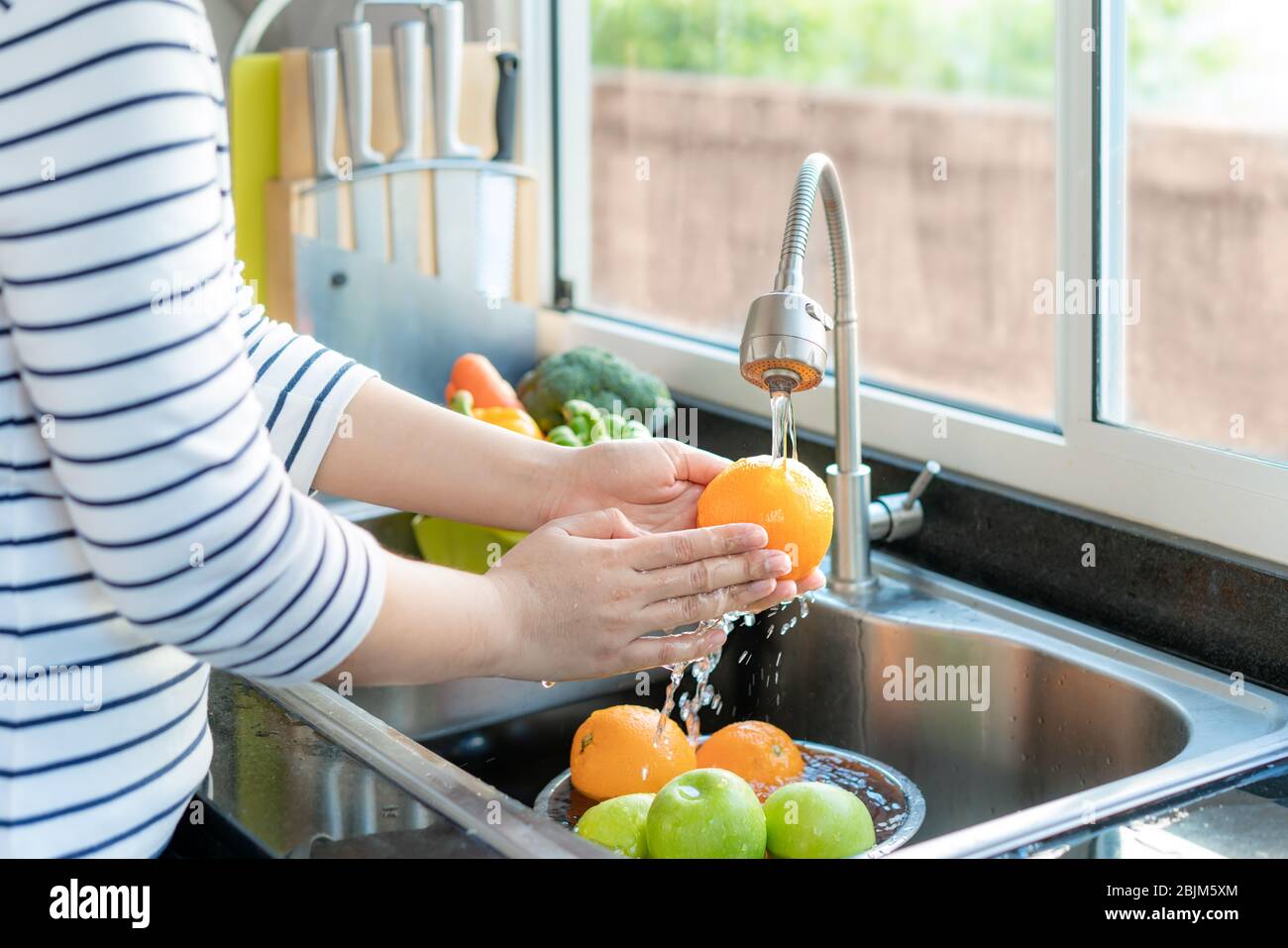 Femme asiatique en bonne santé laver une orange et d'autres fruits  au-dessus de l'évier de cuisine et nettoyer un fruit / légume avec de l'eau  pour éliminer les chances de conta Photo