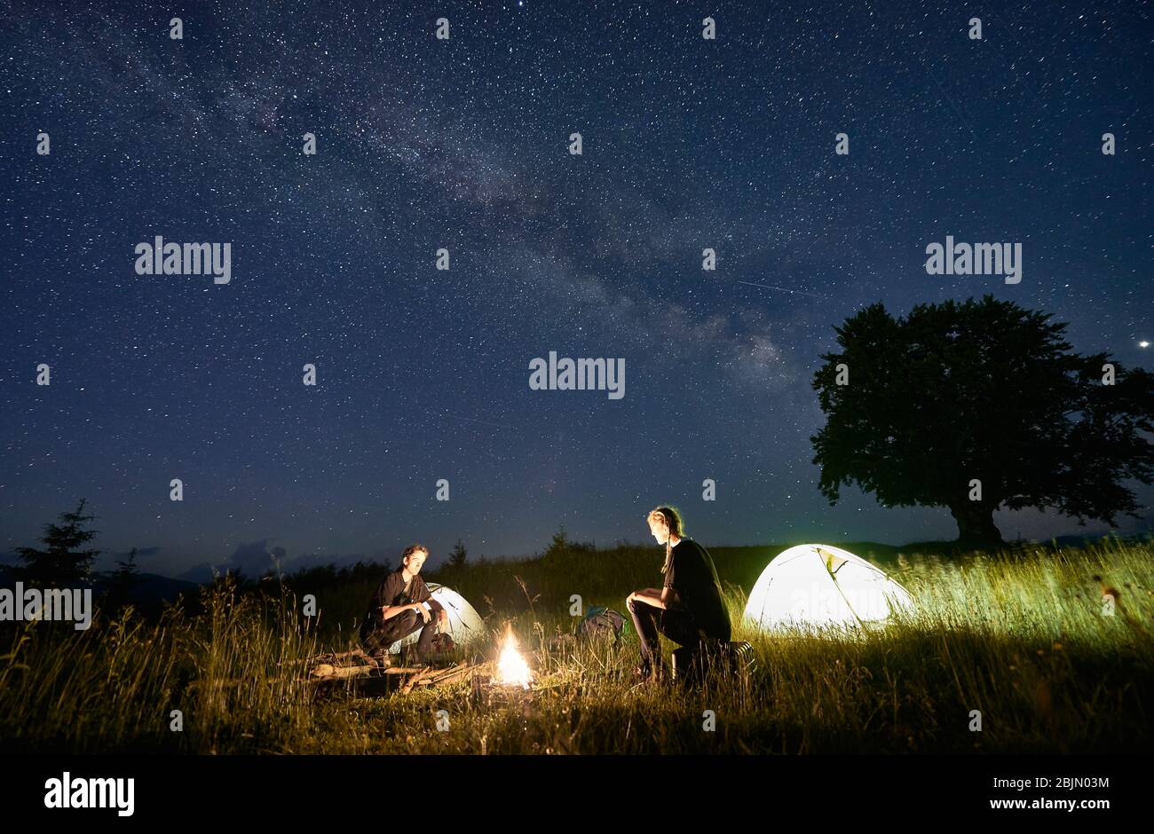 Randonneurs assis devant l'un l'autre sur un banc de grumes, en regardant le feu ensemble à côté du camp et des tentes dans la nuit. Silhouette d'un grand arbre sur fond, voie lactée et ciel étoilé au-dessus d'eux Banque D'Images