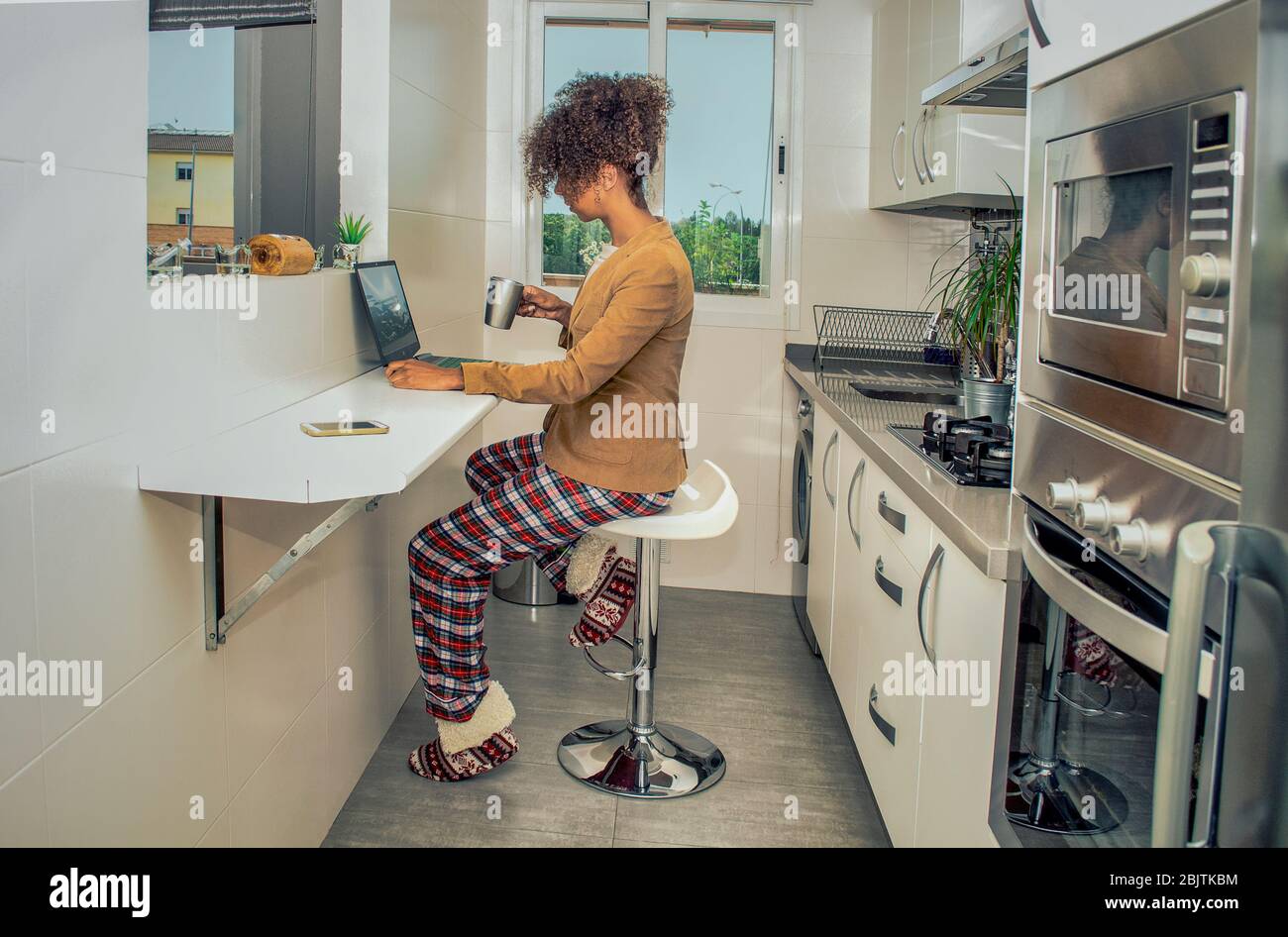 Vue latérale d'une femme noire travaillant à la maison pendant la quarantaine de Coronavirus. Conférence téléphonique dans la cuisine portant pyjama et veste. Banque D'Images