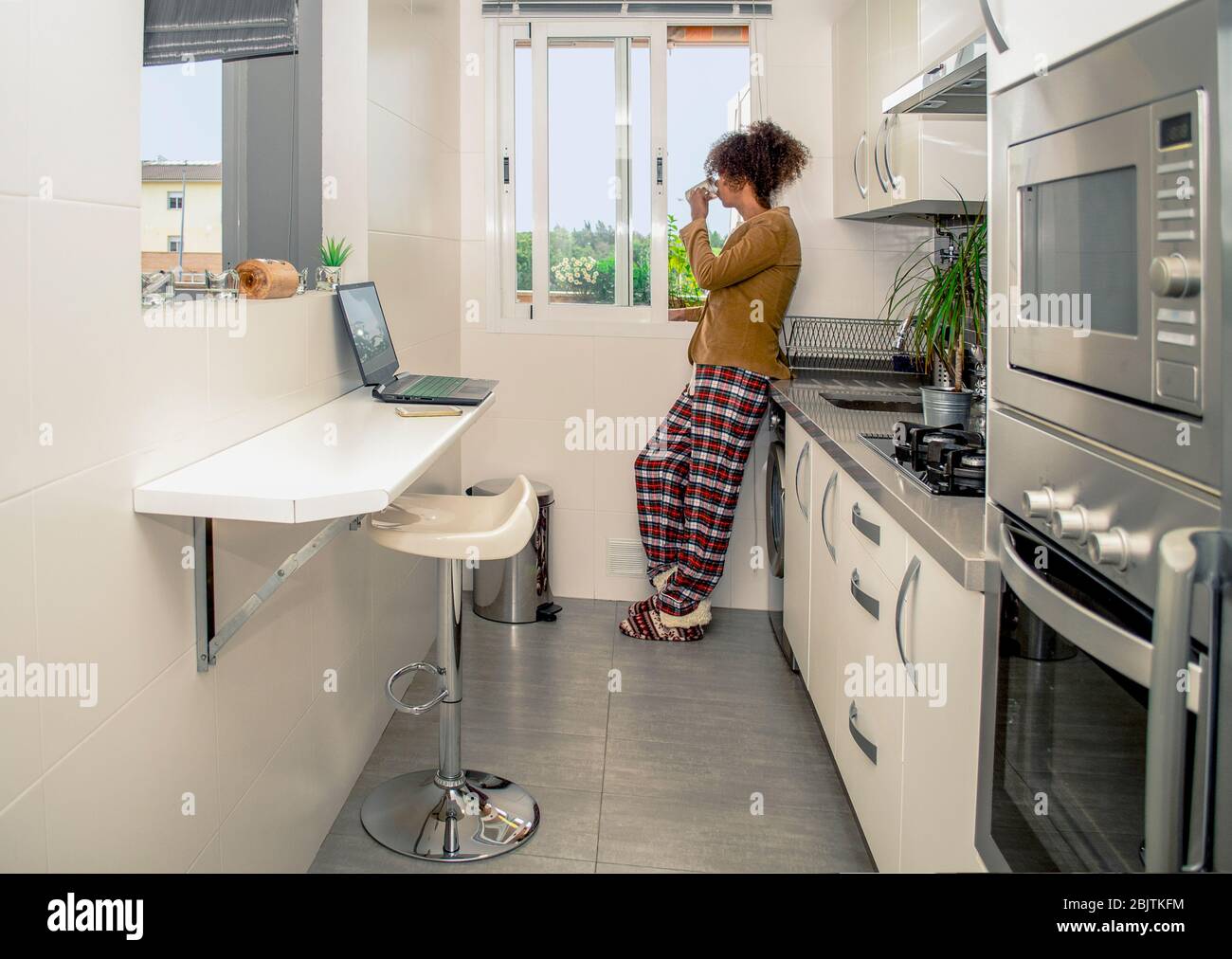Vue latérale d'une femme noire travaillant à la maison pendant la quarantaine de Coronavirus. Conférence téléphonique dans la cuisine portant pyjama et veste. Pause petit déjeuner Banque D'Images