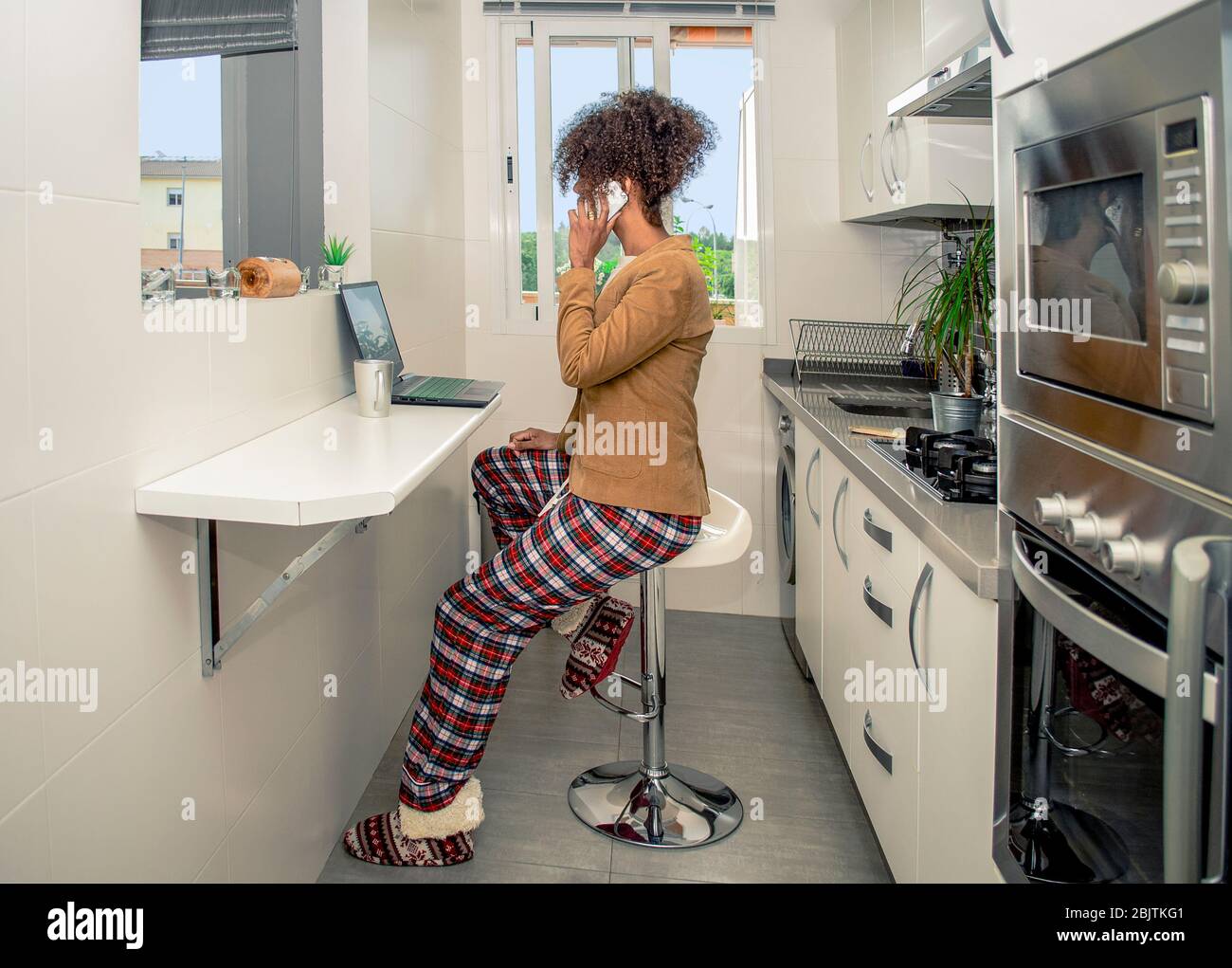 Vue latérale d'une femme noire travaillant à la maison pendant la quarantaine de Coronavirus et tenant un smartphone. Conférence téléphonique dans la cuisine portant pyjama et Banque D'Images