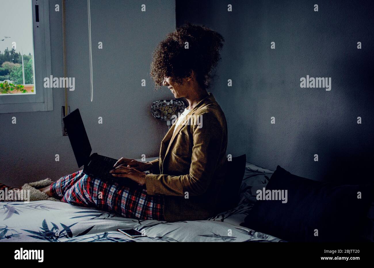 Vue latérale d'une femme noire travaillant à la maison pendant la quarantaine de Coronavirus. Conférence téléphonique dans la chambre avec pyjama et veste. PH cinématographique sombre Banque D'Images