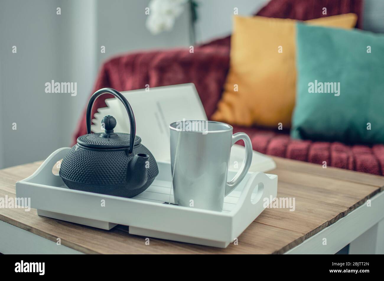 Gros plan et mise au point sélective sur un plateau avec une théière et une tasse sur une table en bois. Couverture bordeaux, coussins jaunes et verts sur un canapé gris dans le bl Banque D'Images