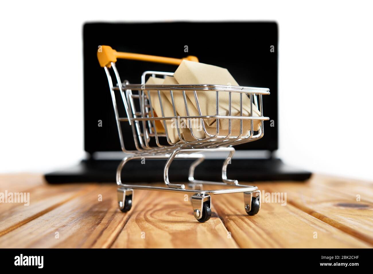Mini supermarché panier plein de boîtes d'expédition se tenir sur une table en bois contre un ordinateur portable noir flou sur fond blanc. Banque D'Images