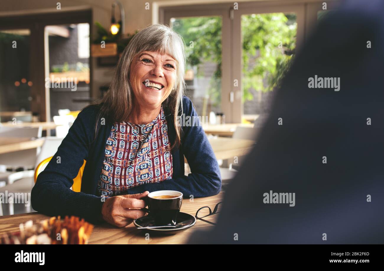 Une femme gaie et senior assise à la table dans un café et parlant avec un homme assis autour. Heureuse femme mûre au café-restaurant. Banque D'Images