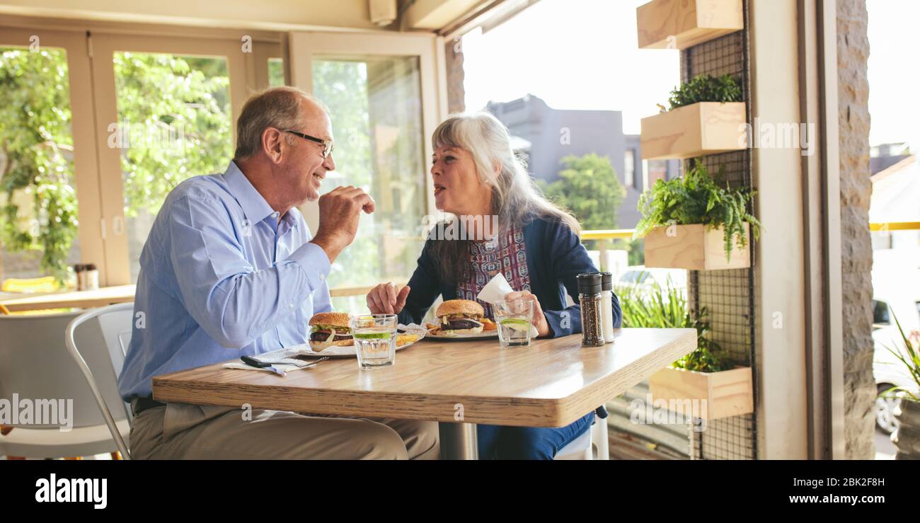 Couple senior qui aime manger du hamburger ensemble dans un café. Homme et femme souriants mangeant un hamburger dans un restaurant. Banque D'Images