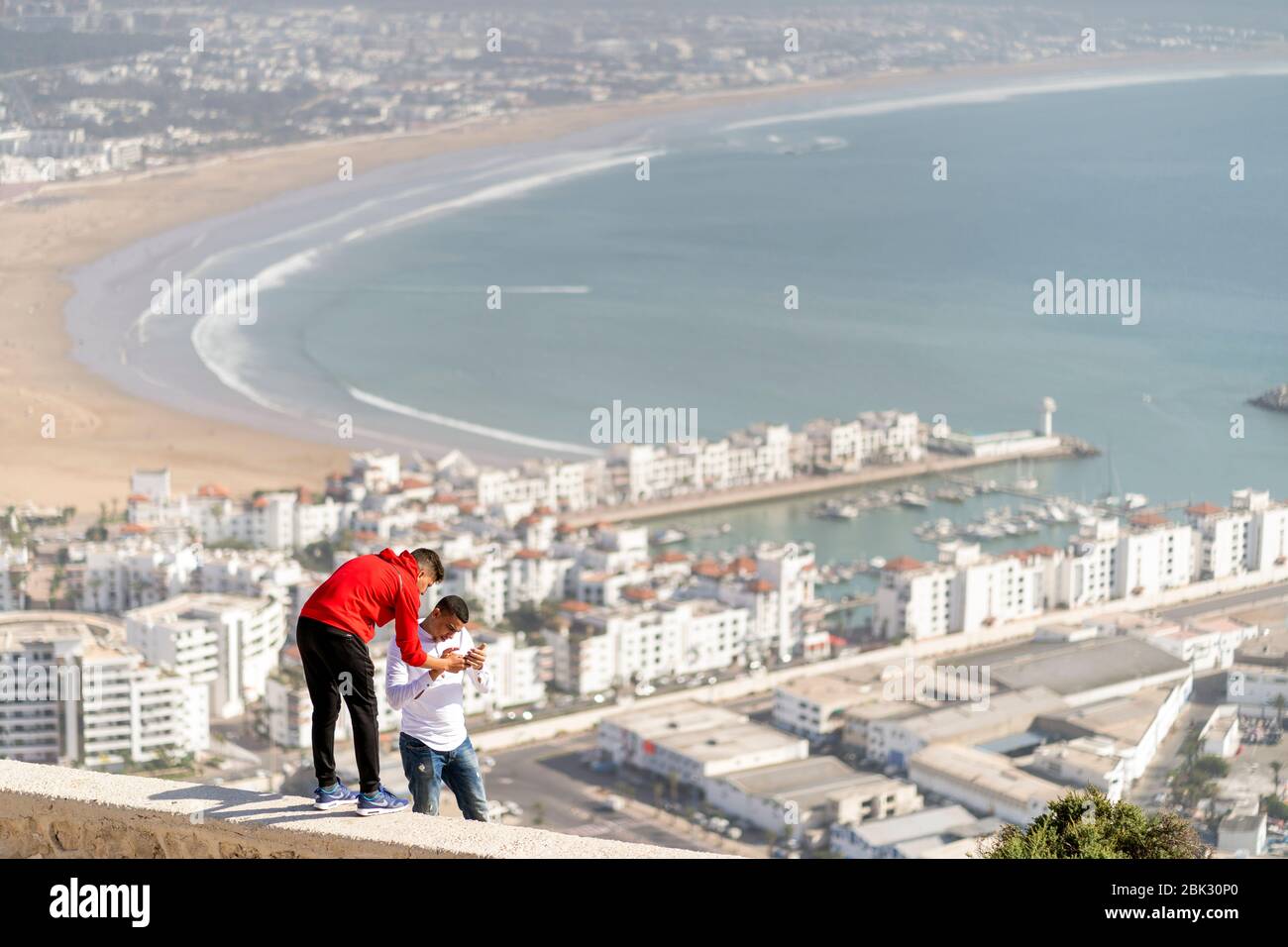Agadir, Maroc - 20 mars 2020: Deux jeunes hommes arabes occupés avec un téléphone mobile sur un beau panorama d'Agadir comme arrière-plan Banque D'Images