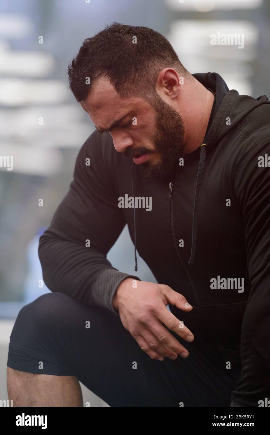 motivation sportive concept de jeune homme barbu fort athlète dans les vêtements de sport sweat à capuche penchant sur le genou pendant la pause dans l'entraînement Banque D'Images