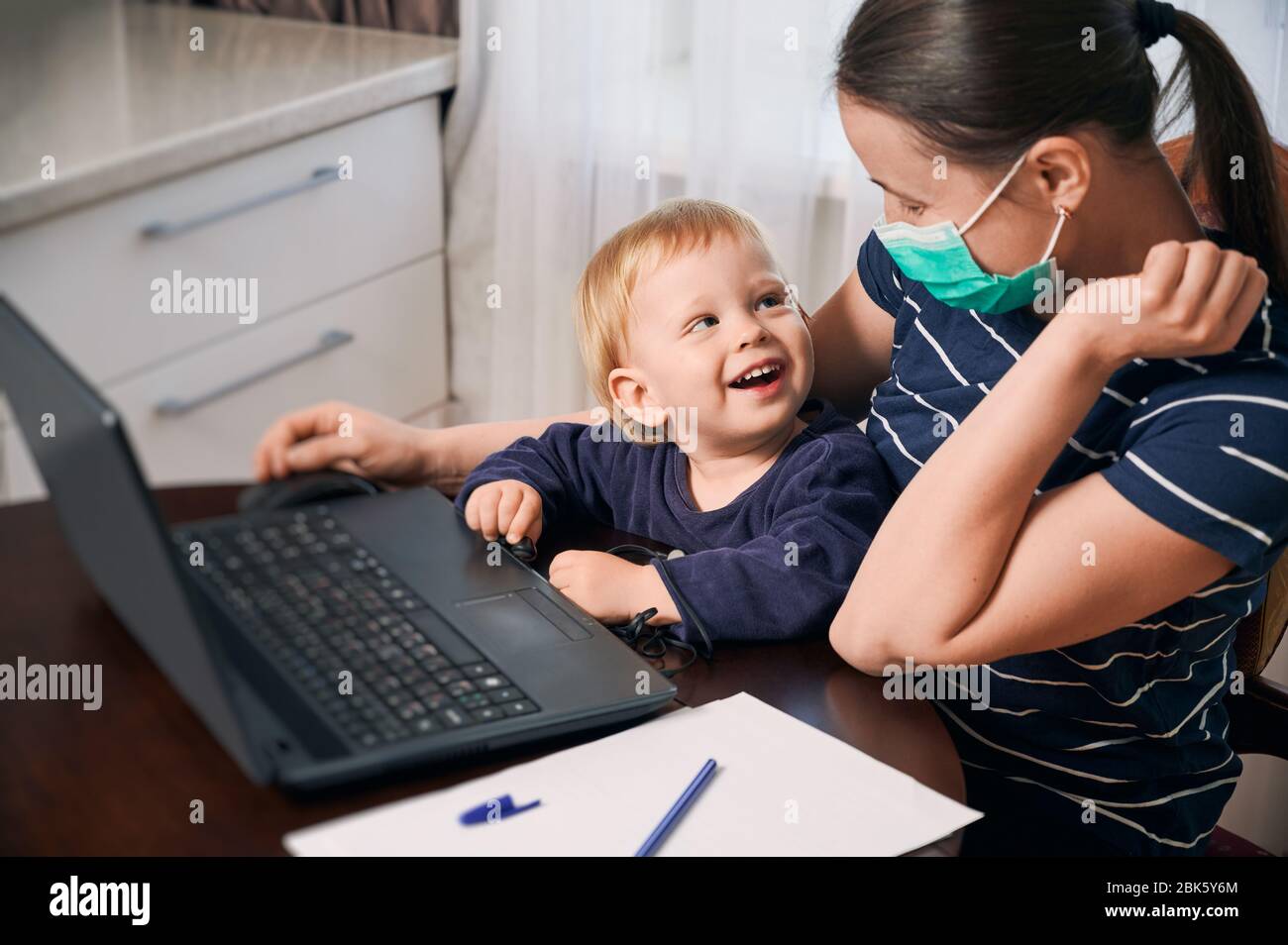 Auto-isolement pour protéger votre famille contre l'infection à coronavirus. Mère portant un masque jouant avec son adorable bébé souriant sur ses tours à pc. Passer plus de temps avec la famille Banque D'Images