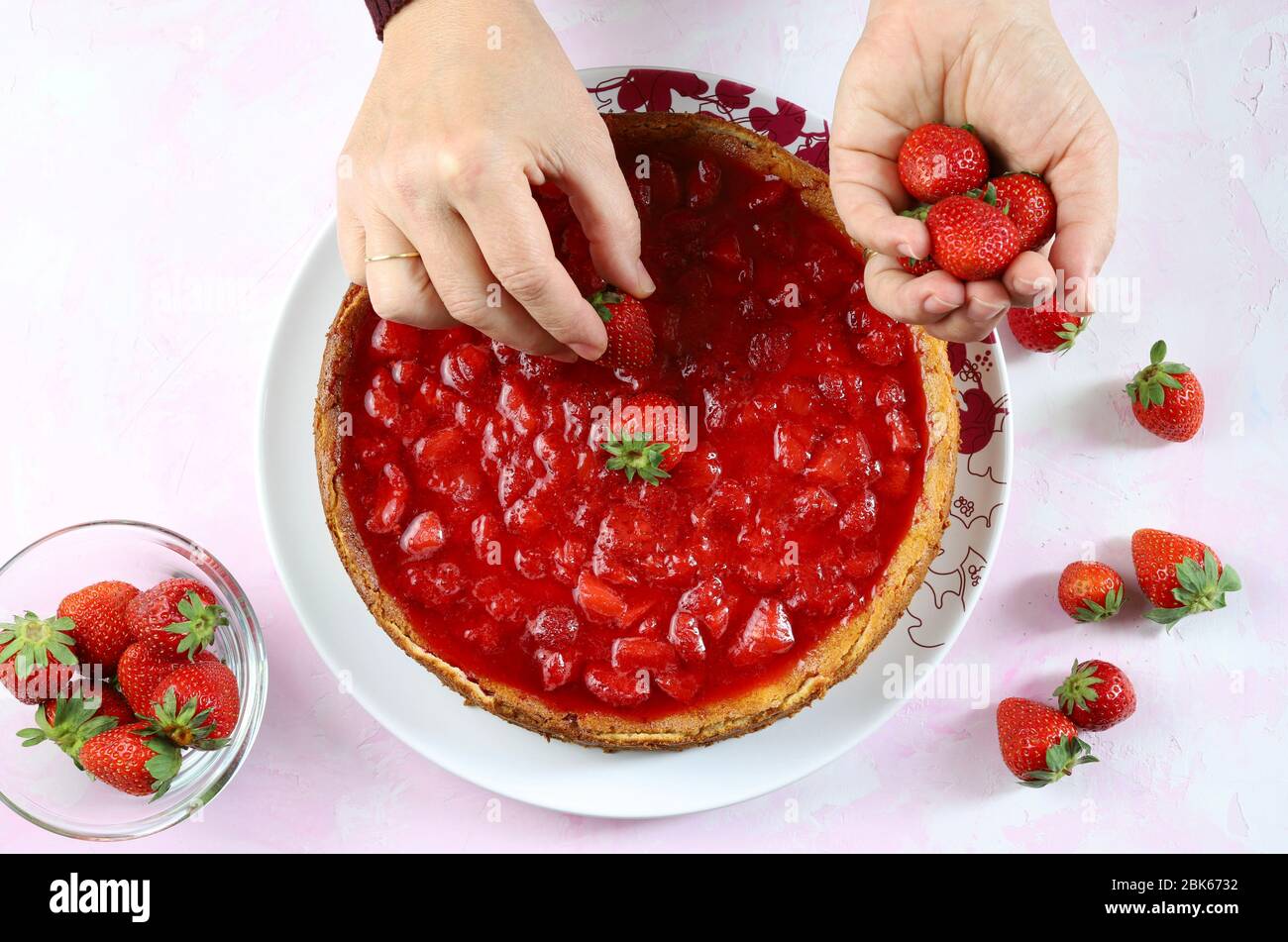 Fraisier cheesecake making.woman décorant à la main avec des fraises fraîches un cheesecake. Fond blanc avec des nuances de rose.vue du dessus.concept Sweet. Banque D'Images