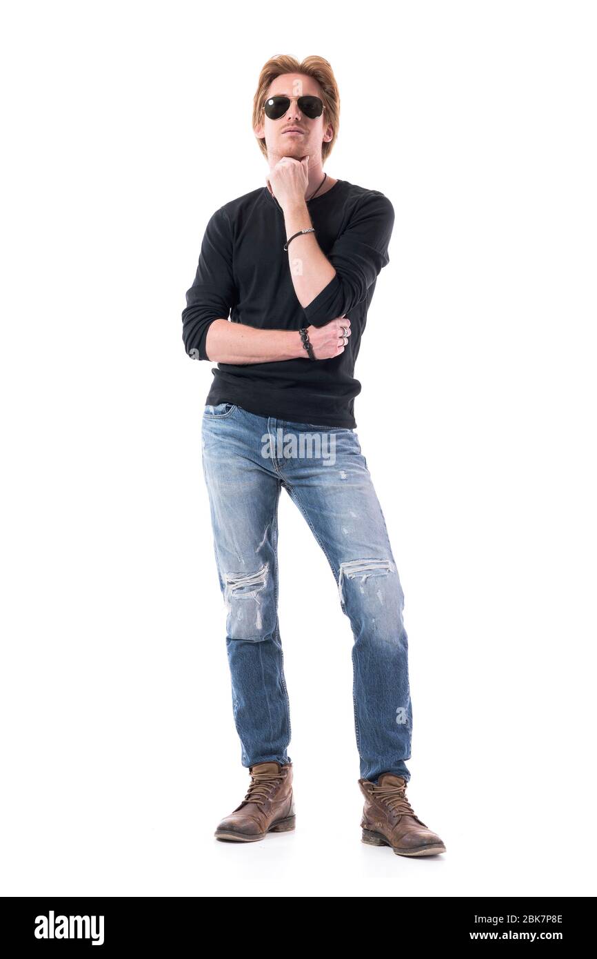 Un jeune homme sérieux et confiant avec des lunettes de soleil se tenant à la main sur le menton regardant et évaluant. Corps entier isolé sur fond blanc. Banque D'Images