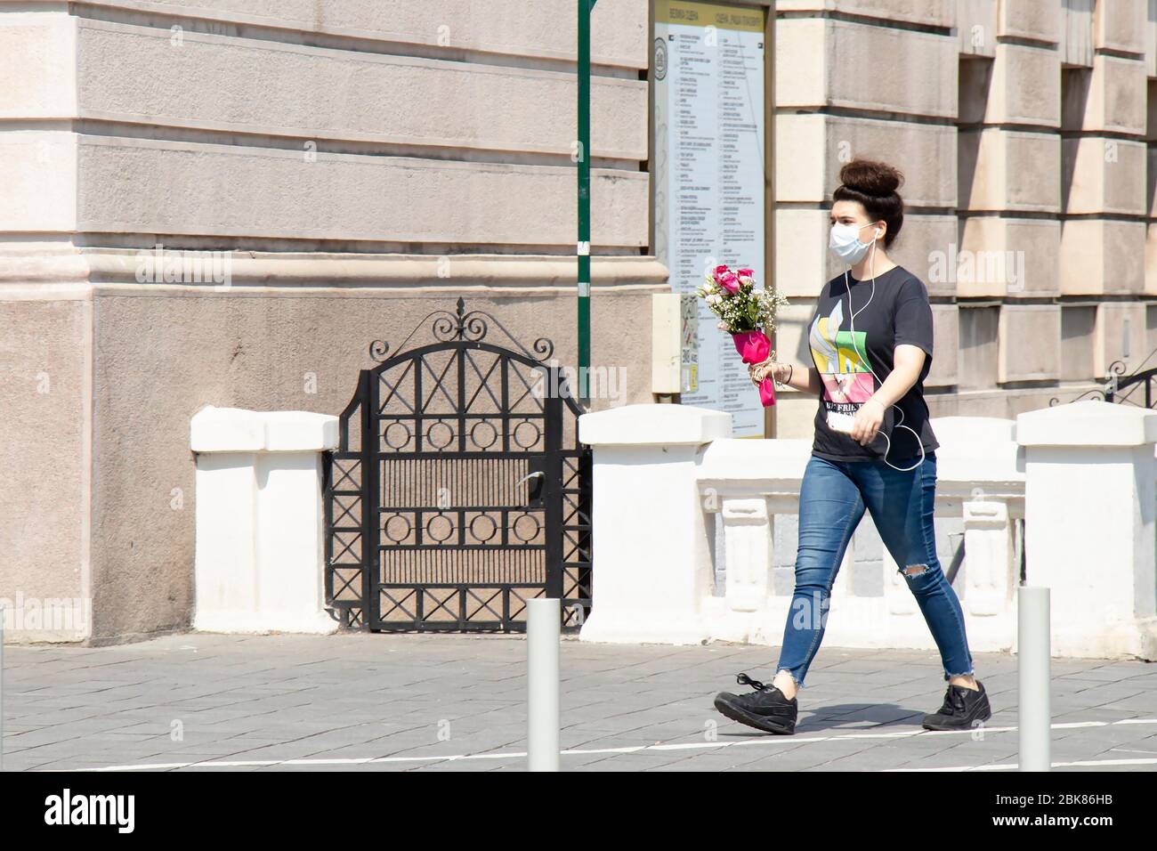 Belgrade, Serbie - 24 avril 2020: Une adolescente portant un masque de visage marchant seule rue de ville par un jour ensoleillé tout en tenant un bouquet de roses dans ses mains Banque D'Images