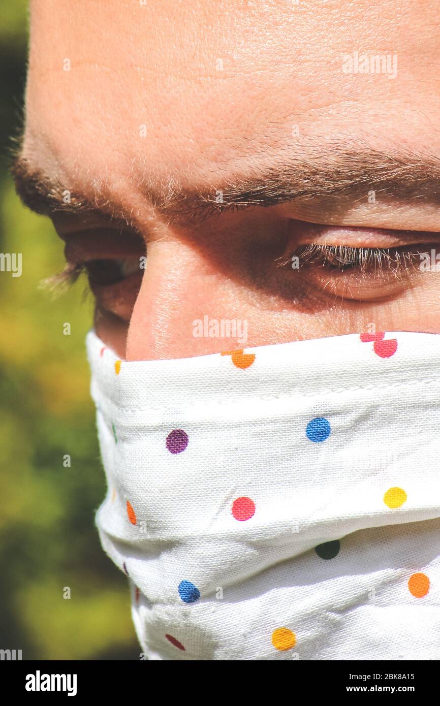 Jeune homme caucasien avec un masque de visage fait main en tissu pendant la pandémie de coronavirus. Coronavirus, éclosion de COVID-19. Concept de protection antivirus. Arrière-plan flou. Détail visage et yeux. Photo verticale. Banque D'Images