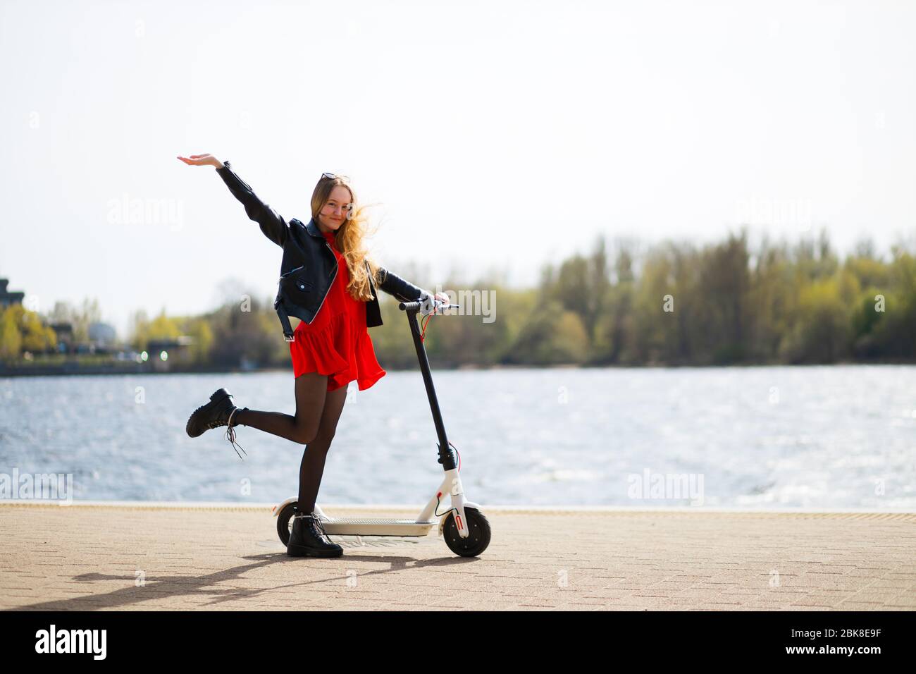 Une jeune fille blonde heureuse et positive lève la main et la jambe près du scooter électrique Banque D'Images