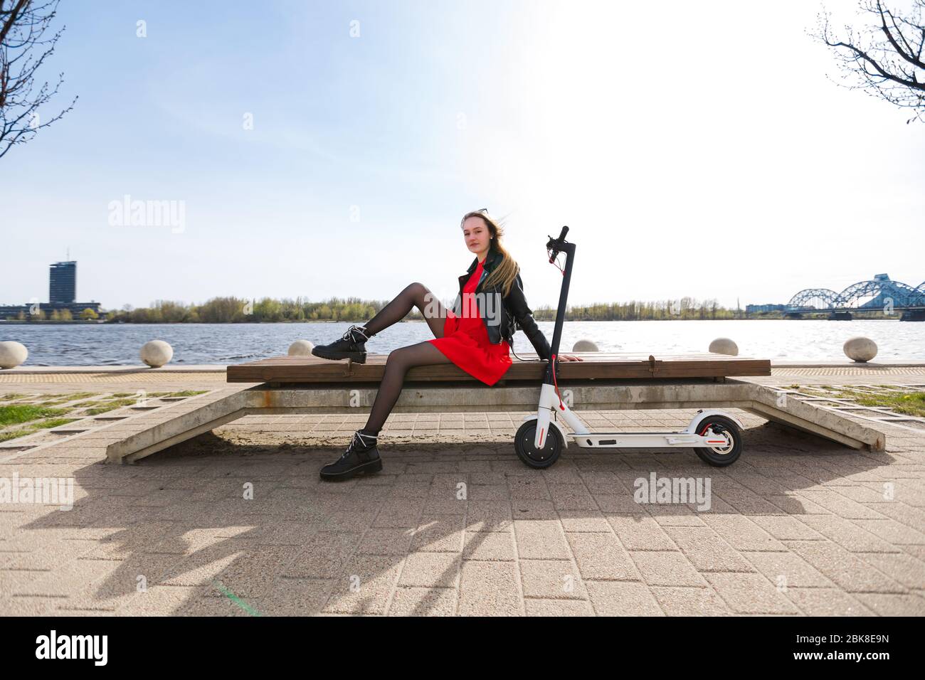 Belle jeune femme blonde est assise sur un banc avec scooter électrique blanc Banque D'Images