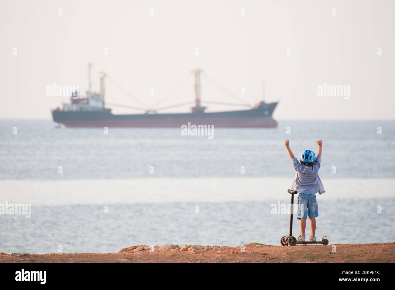 joyeux petit enfant en casque avec scooter sur la plage de la mer avec cargo à l'horizon Banque D'Images