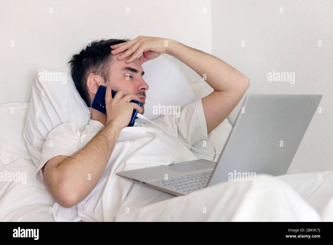 Un homme adulte est allongé dans un lit d'hôpital avec un ordinateur portable tenant sa tête. Concept de travail à distance pendant la pandémie du coronavirus Banque D'Images