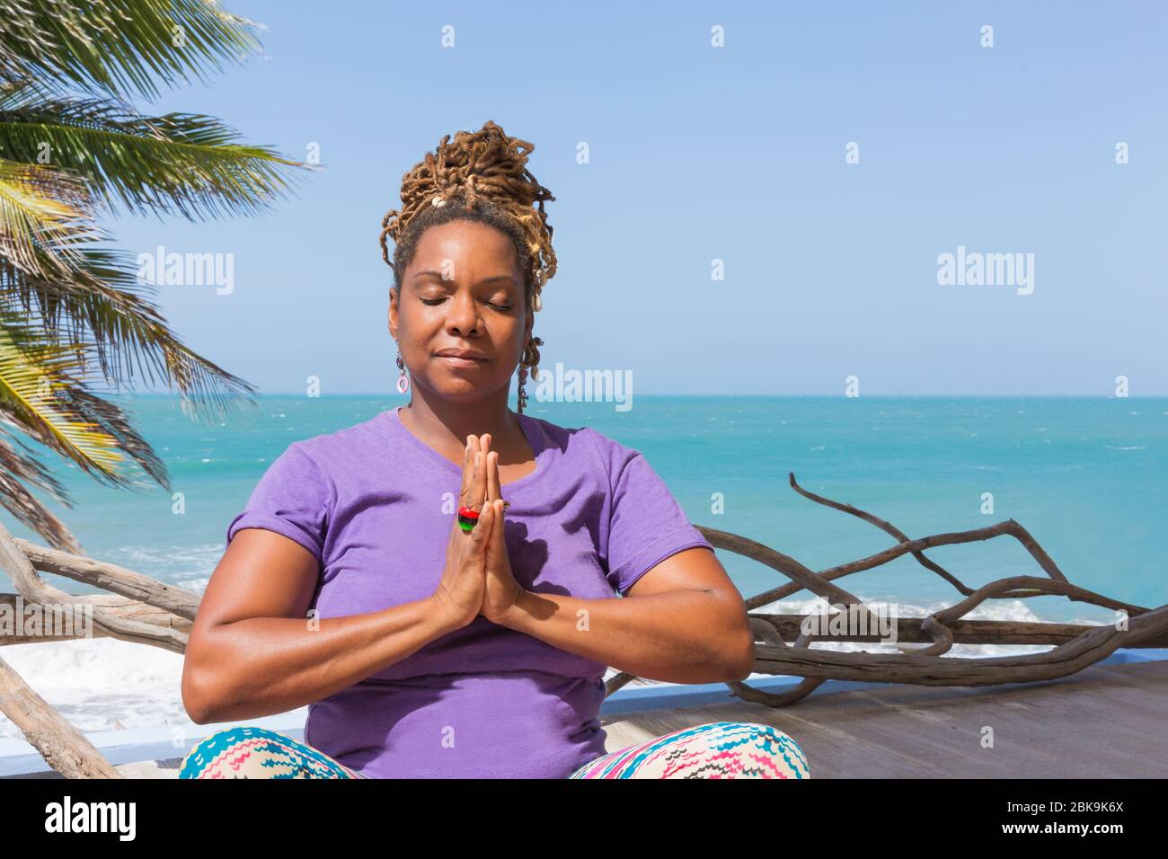 Belle femme noire, africaine ou caribéenne pratiquant le bien-être et pose de yoga dans le complexe ou l'hôtel à l'extérieur surplombant la plage et l'océan magnifiques. Banque D'Images