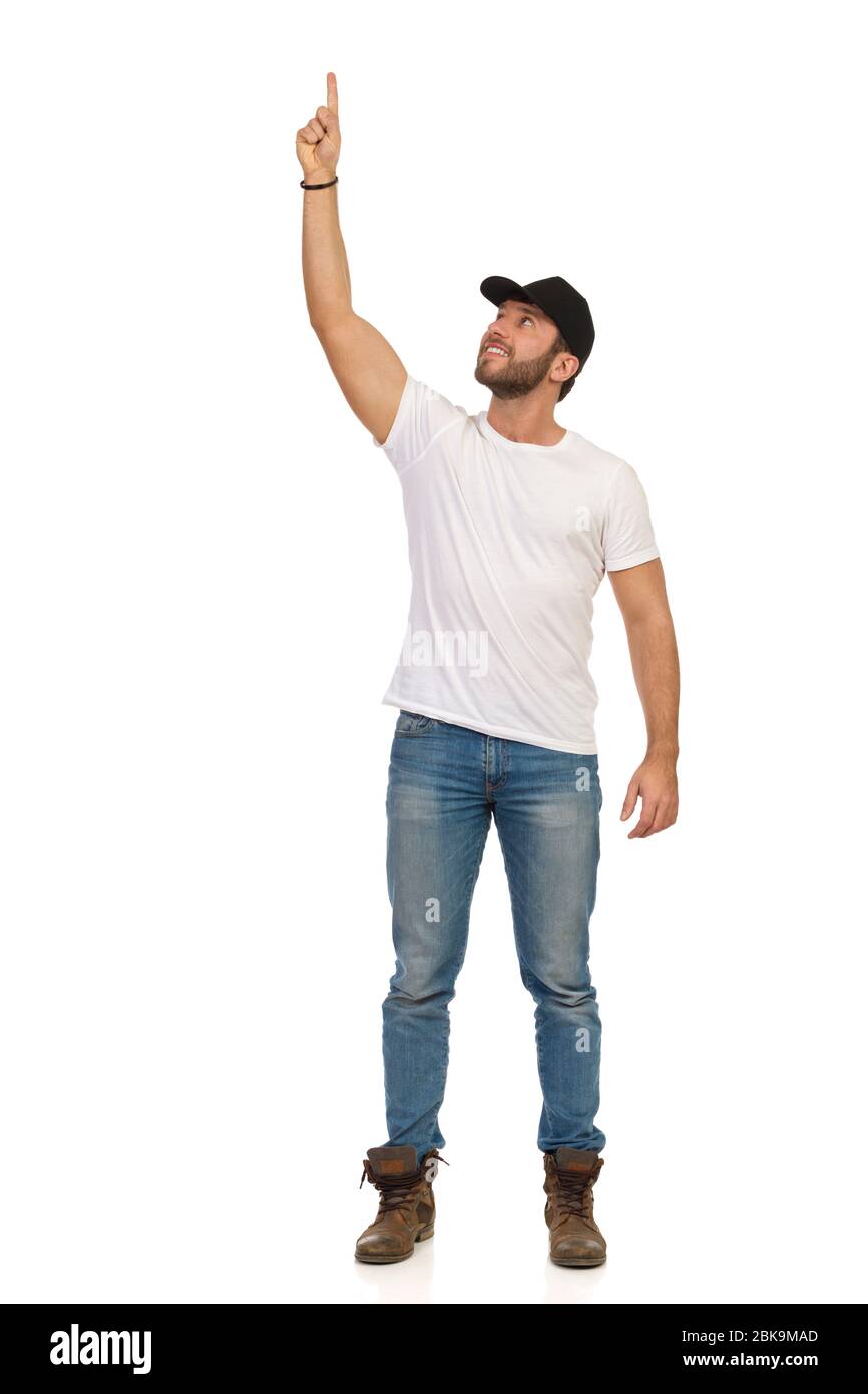 Homme en Jean, t-shirt blanc et casquette noire est debout avec bras levé, pointant vers le haut et regardant. Studio complet tourné isolé sur blanc. Banque D'Images