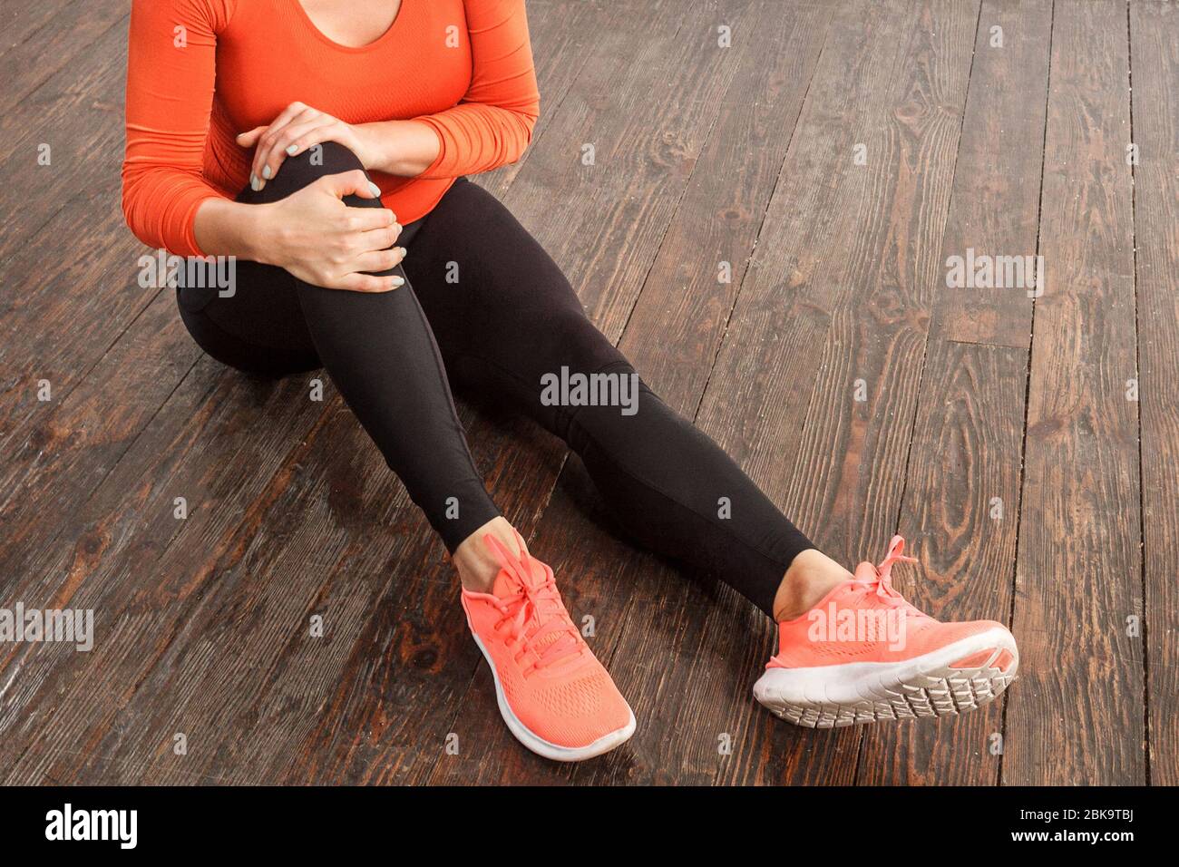 Femme dans des vêtements de sport serrés tenant le genou douloureux assis sur le sol à la salle de gym à la maison, souffrant de la tension musculaire, des ligaments de pluie ou des blessures articulaires, pro de santé Banque D'Images
