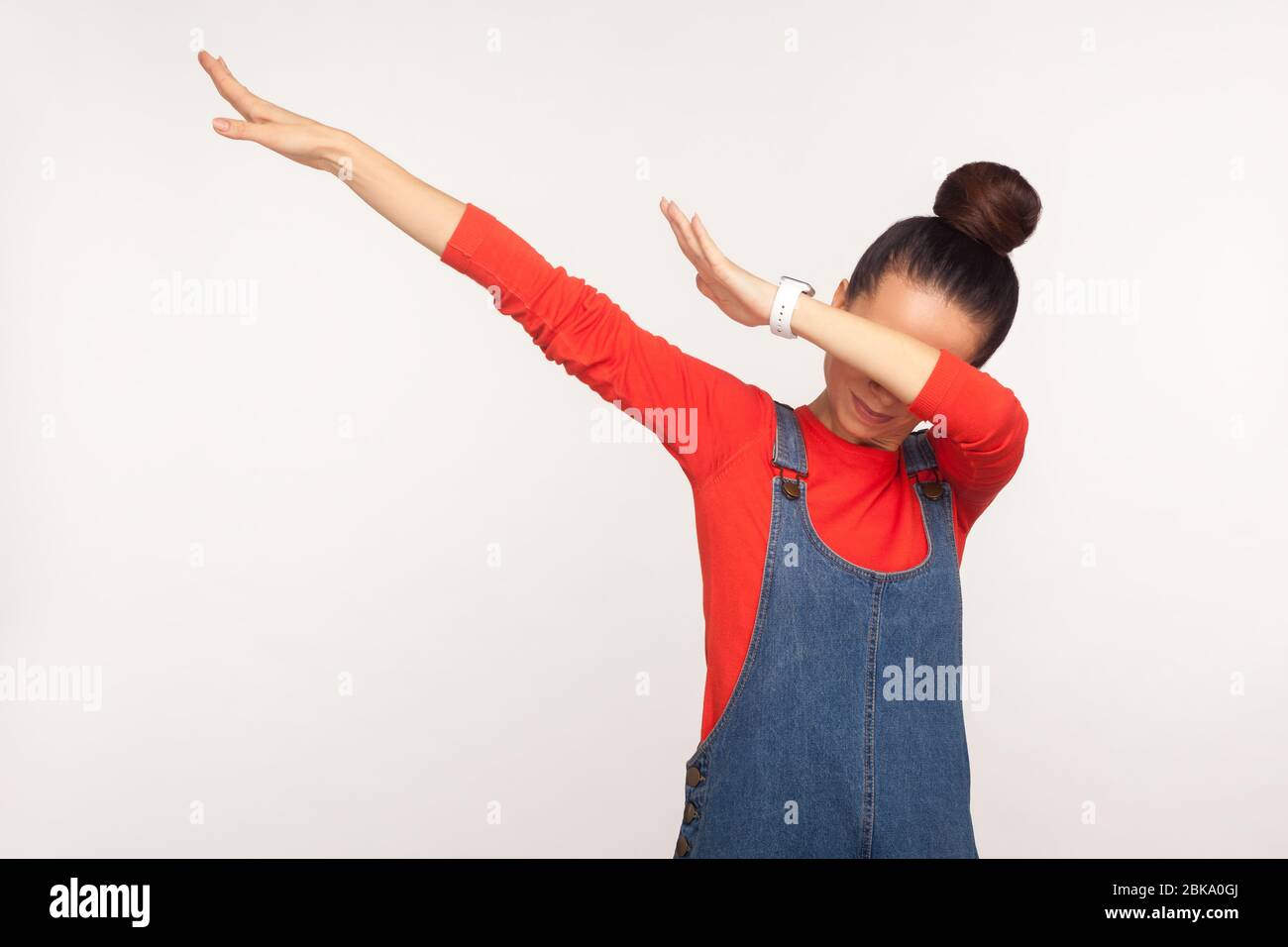 Victoire, geste de succès. Portrait d'une fille élégante avec un pain aux cheveux dans des combinaisons en denim montrant une posture de danse dab, des souvenirs Internet célèbres, exécutant un tr de dabbing Banque D'Images