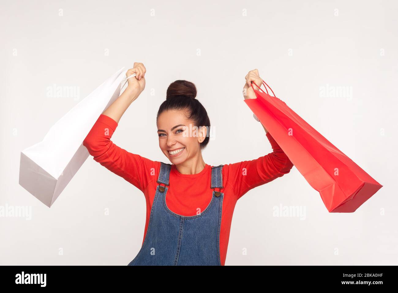 Des achats géniaux ! Portrait d'une jeune fille joyeuse et élégante avec un pain aux cheveux dans des salopettes en denim, en levant des paquets et en regardant un appareil photo avec un sourire excité, enj Banque D'Images