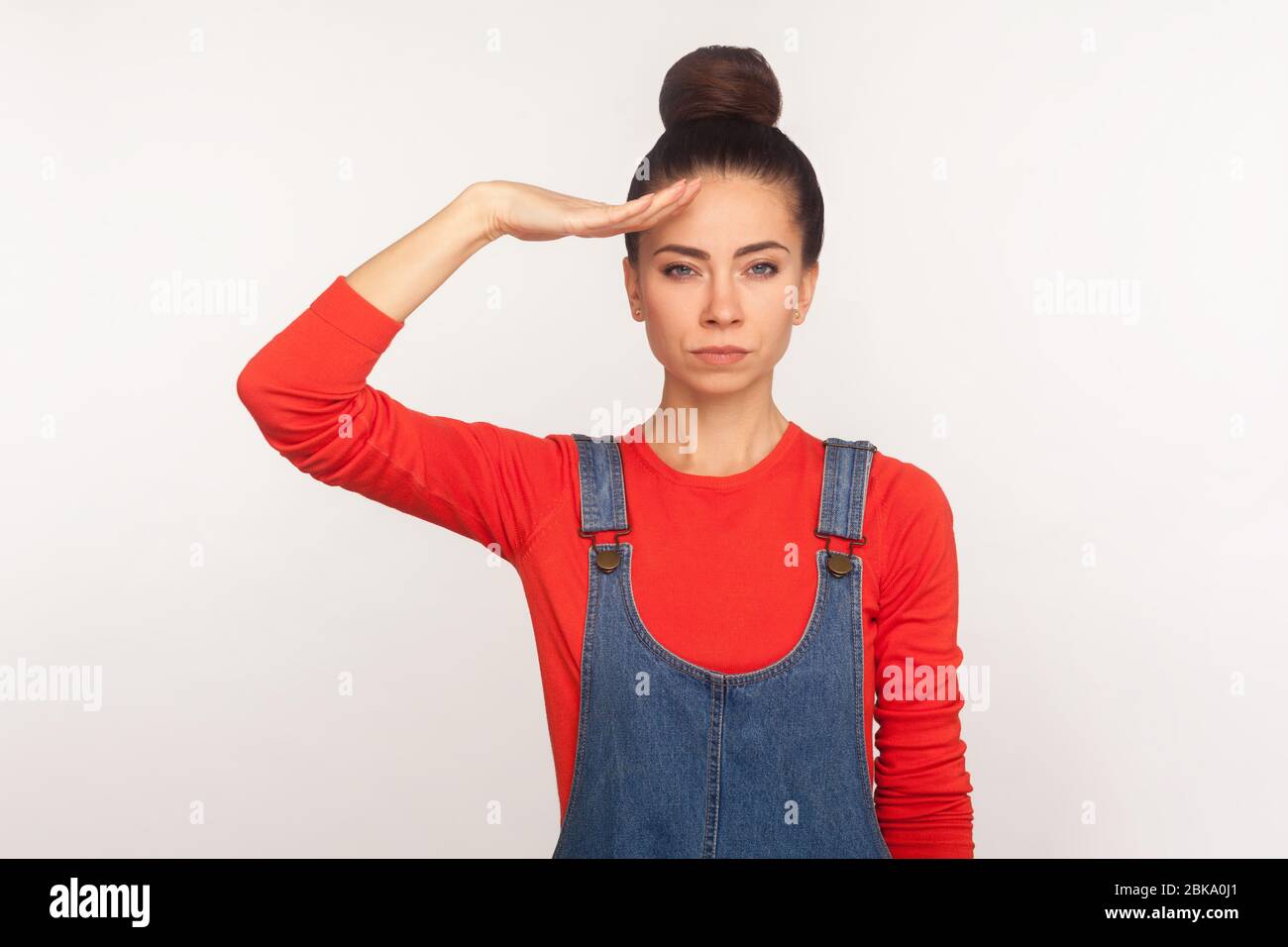 Oui monsieur! Portrait d'une fille responsable stricte avec un pain de cheveux dans des salopettes en denim donnant saluant l'écoute de l'ordre avec une expression sérieuse et attentive, follo Banque D'Images