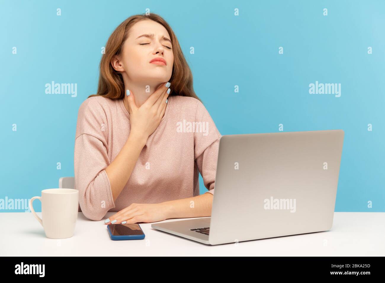 Jeune femme malade de la grippe assis sur le lieu de travail avec ordinateur portable et tenant le cou douloureux, souffrant de maux de gorge, des amygdales enflammées, des symptômes de la grippe. Banque D'Images