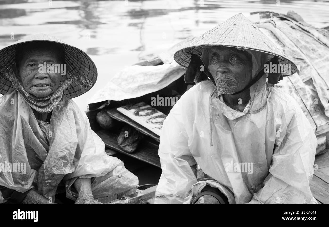 Hoi an Vietnam 17 octobre 2020; couple vietnamien senior assis leur petit bateau dans la rivière abritant de la pluie dans des chapeaux coniques et des couvertures en plastique dans Banque D'Images