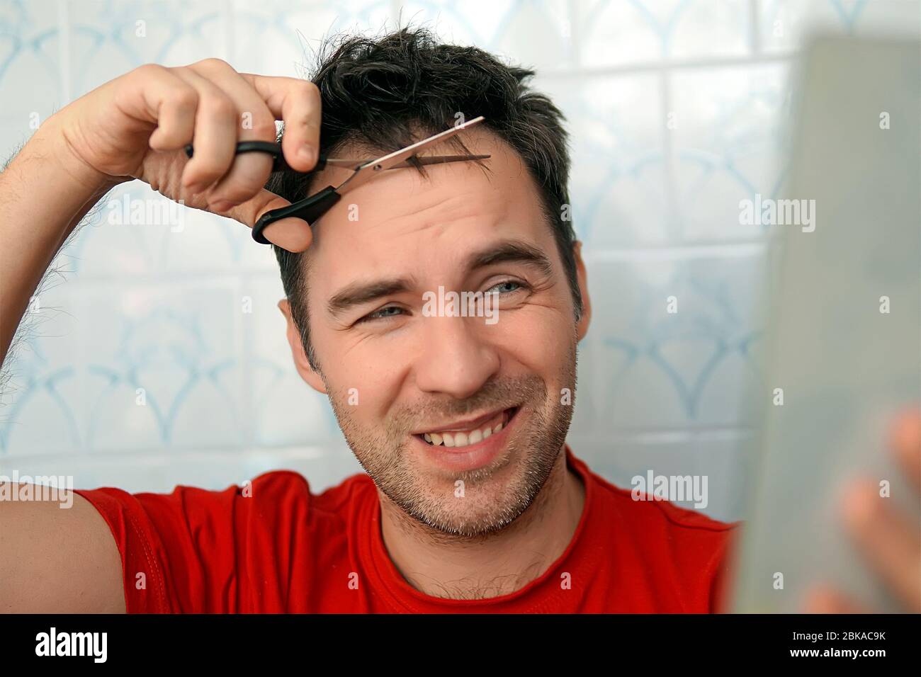 Beau homme coupant ses propres cheveux avec une paire de ciseaux et regarde dans le miroir. Autosoins dans les conditions de quarantaine globale et fermé coiffeurs an Banque D'Images