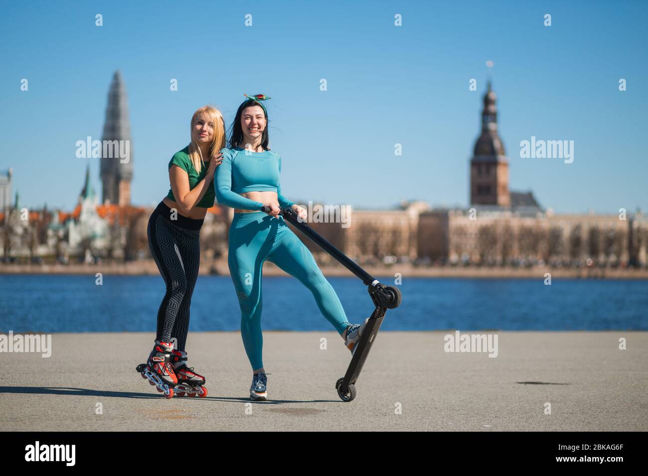 Les sœurs blonde et brunette joyeuses s'amusent avec des patins à roulettes et un scooter électrique sur la promenade de la ville Banque D'Images