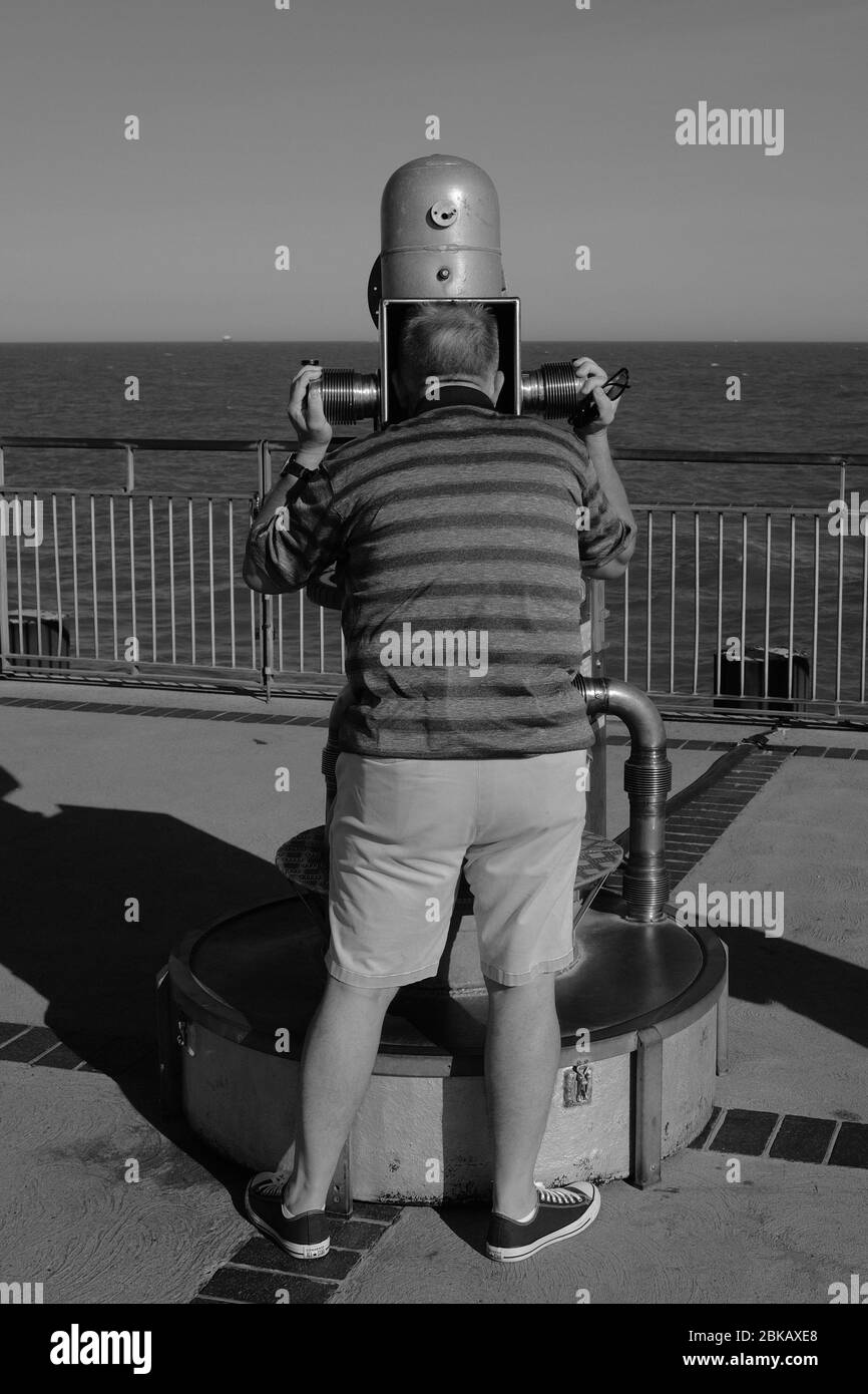 Homme regardant trop la mer à travers un télescope étrange depuis Southwold Pier suffok uk en noir et blanc Banque D'Images
