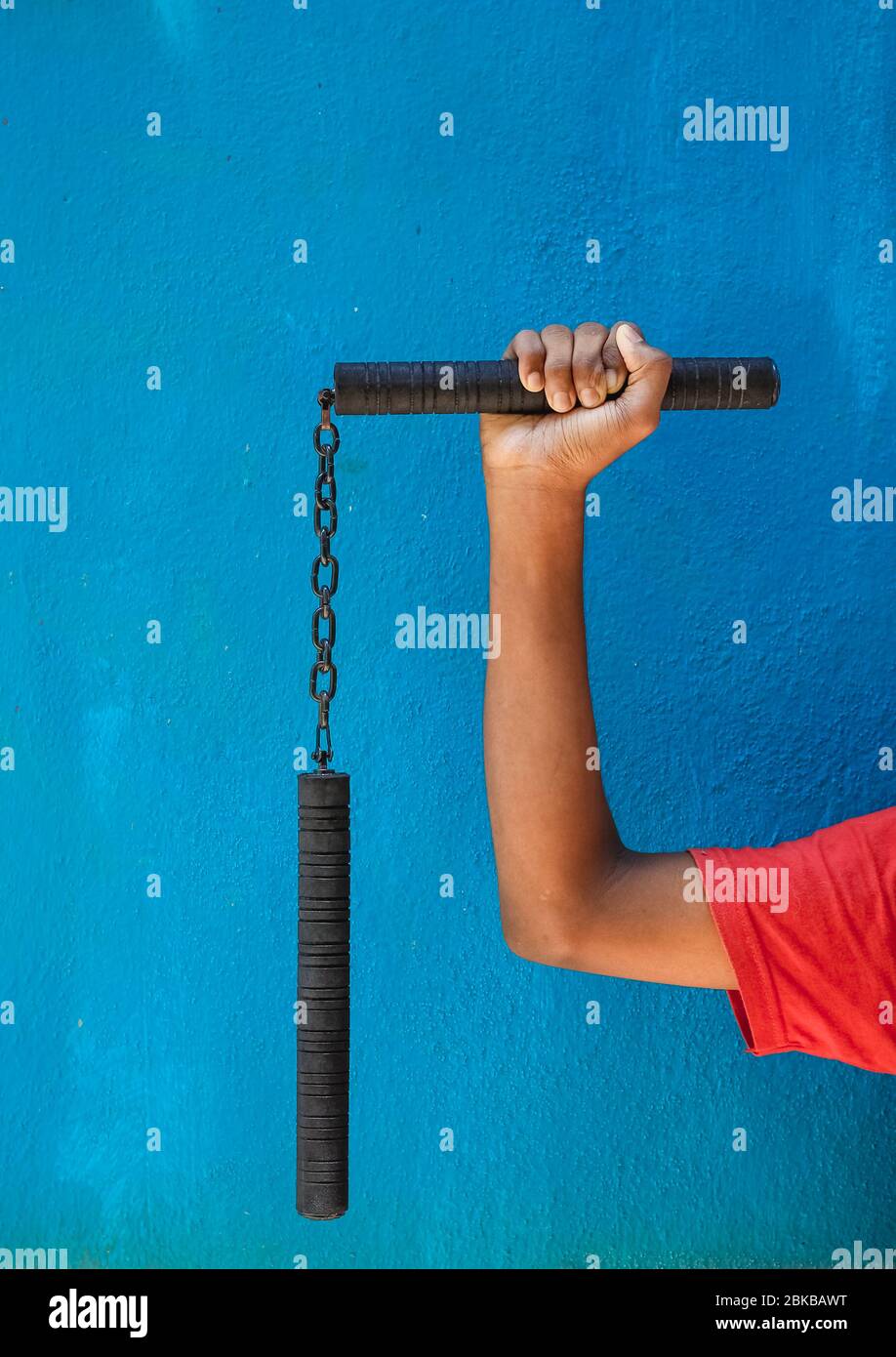 Garçon tenant Nunchaku sur sa main isolé sur fond bleu. Formation nunchaku noir pour débutant. Équipement d'arts martiaux Hot Tactical Gear. Banque D'Images