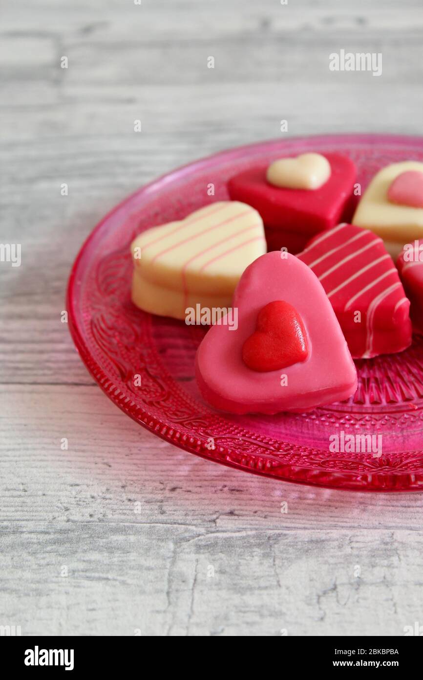 Chocolats colorés en forme de coeur (petits fours) sur une assiette en verre rose avec motif décoratif. Arrière-plan de la table en bois clair. Banque D'Images