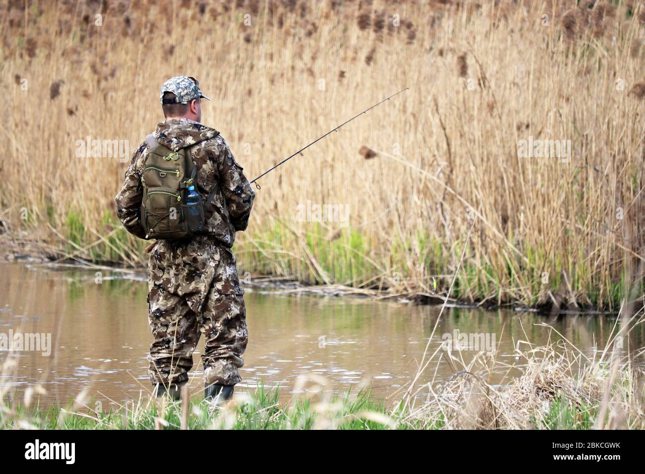 Pêcheur dans des vêtements de chasse debout avec une tige de pêche. Homme à la pêche au printemps sur la côte de la rivière surmonté avec des roseaux Banque D'Images