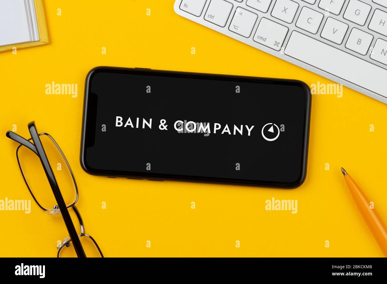 Un smartphone affichant le logo bains & Company repose sur un fond jaune avec un clavier, des lunettes, un stylo et un livre (usage éditorial uniquement). Banque D'Images