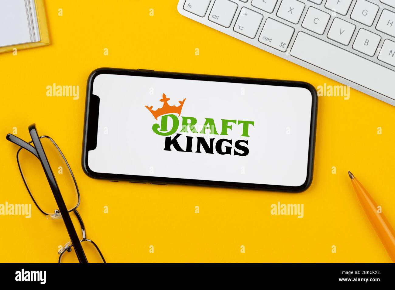 Un smartphone affichant le logo Draft Kings repose sur un fond jaune avec un clavier, des lunettes, un stylo et un livre (usage éditorial uniquement). Banque D'Images