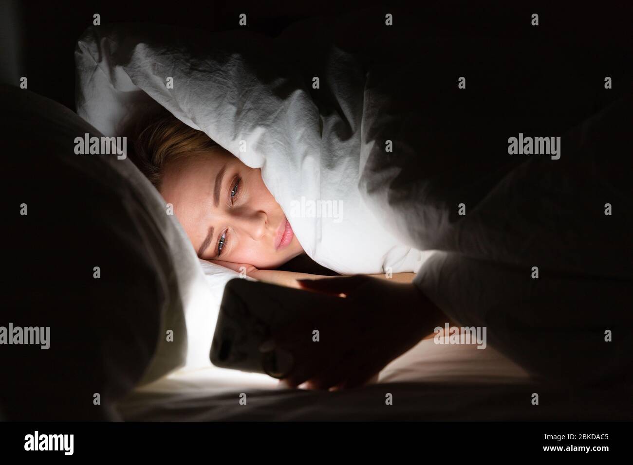 Femme posée au lit sous la couverture en utilisant un smartphone tard dans la nuit, ne peut pas dormir, accro des nouvelles sur Covid-19 ou le défilement des réseaux sociaux. Insomnie, Banque D'Images