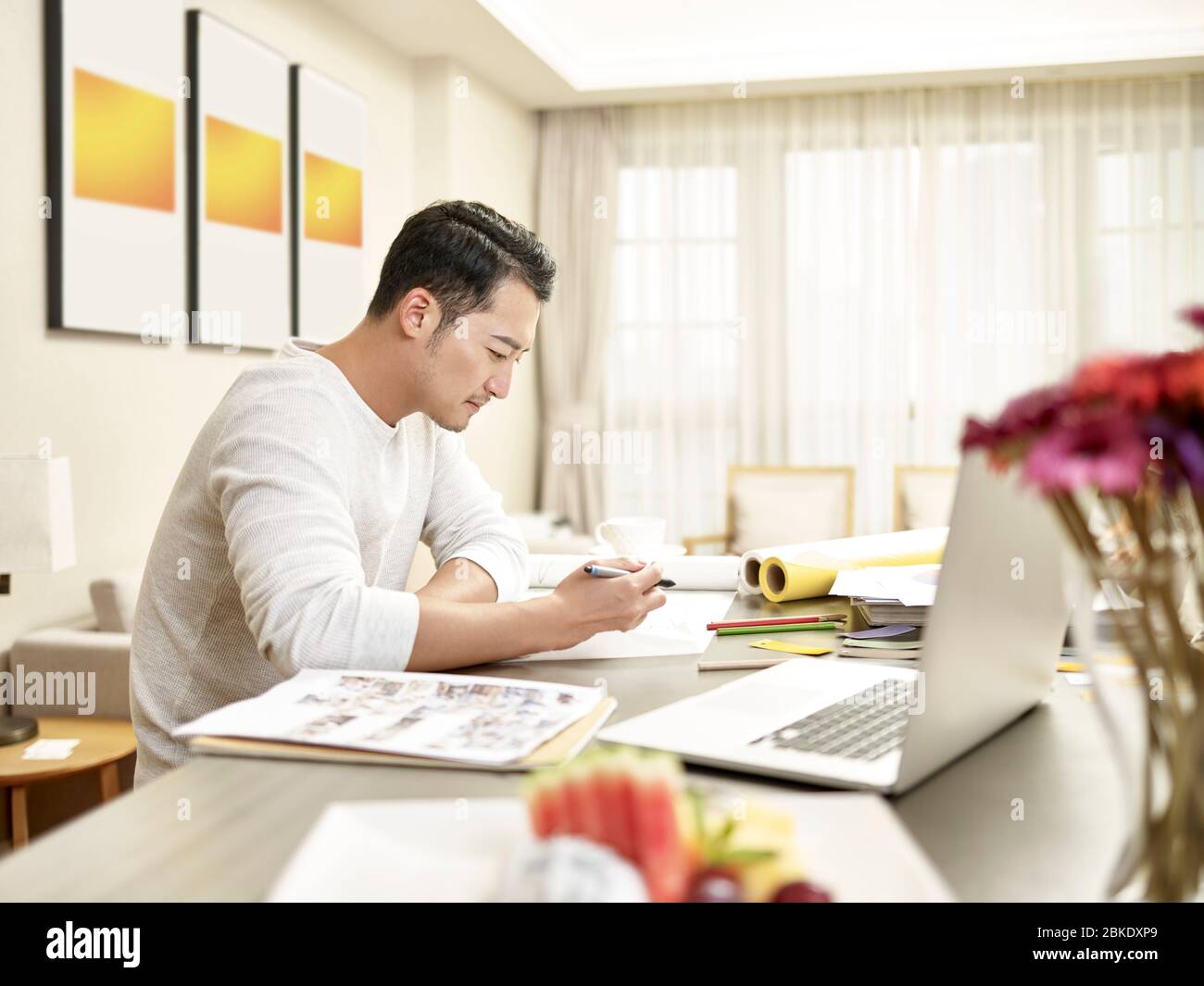 jeune homme asiatique design professionnel assis sur le comptoir de cuisine travaillant à la maison (œuvres d'art en arrière-plan modifiées numériquement) Banque D'Images