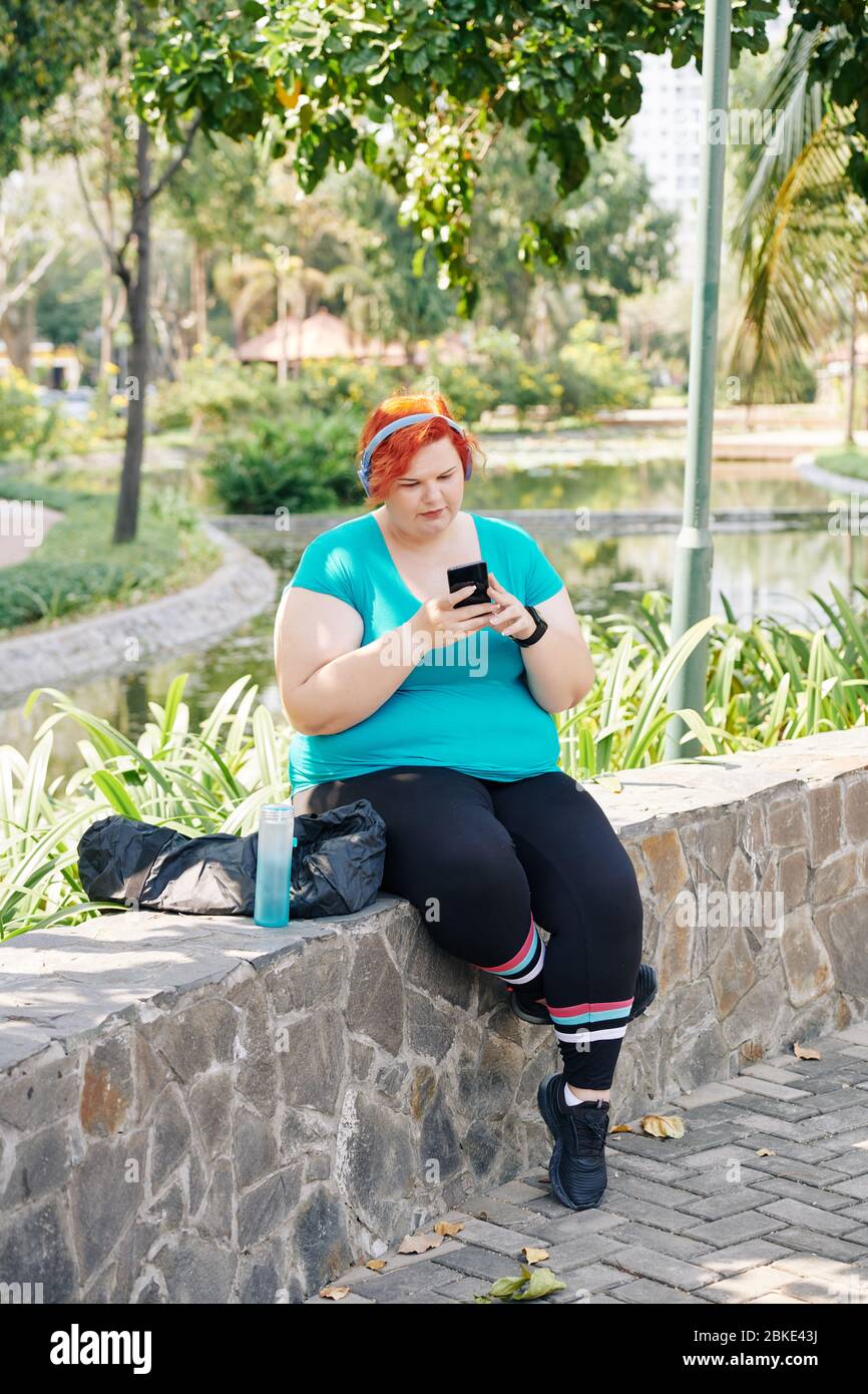 Jeune femme de taille plus se reposant sur un mur de pierre dans le parc et vérifiant les messages et les notifications dans son smartphone Banque D'Images