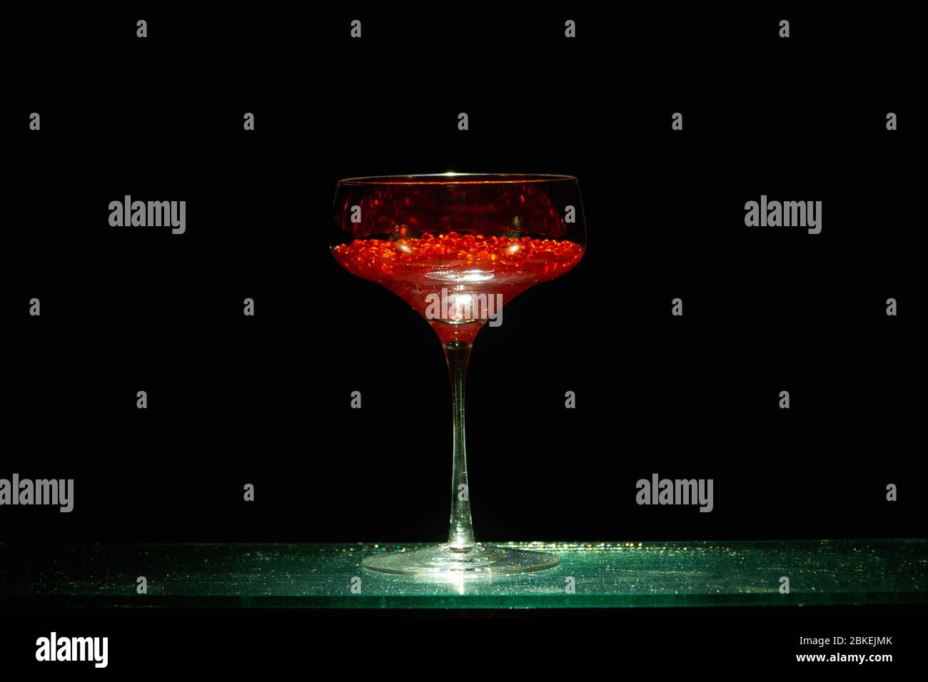 Un seul verre à cocktail de style martini illuminé d'un caviar d'eau rouge de couleur vive dans le style d'un cocktail. Verrerie d'art Banque D'Images