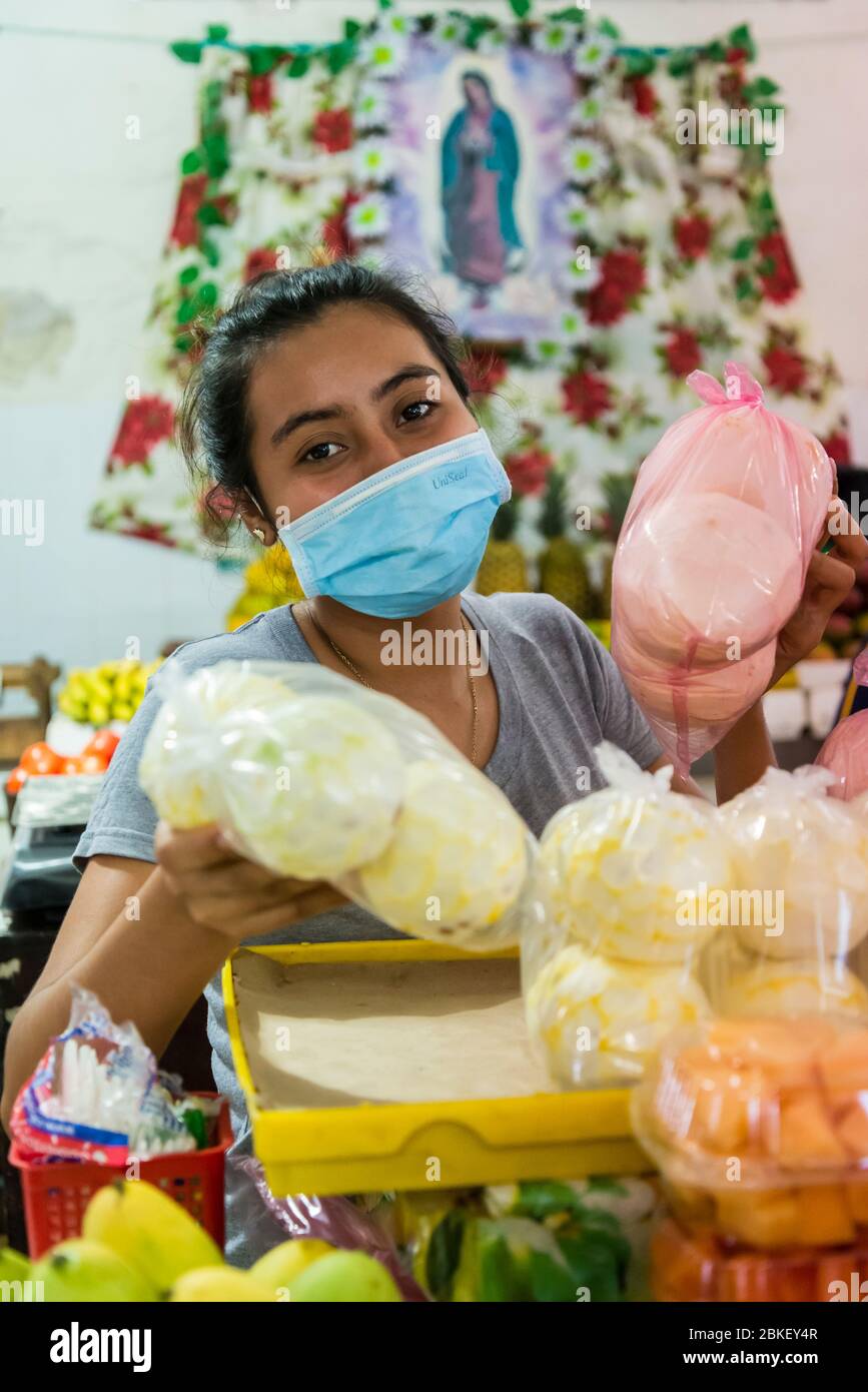 Jeune femme vendeur avec masque chirurgical et photo de Vierge Marie derrière sa vente de nourriture enveloppée dans le plastique au Mercado de Santiago, Merida, y Banque D'Images
