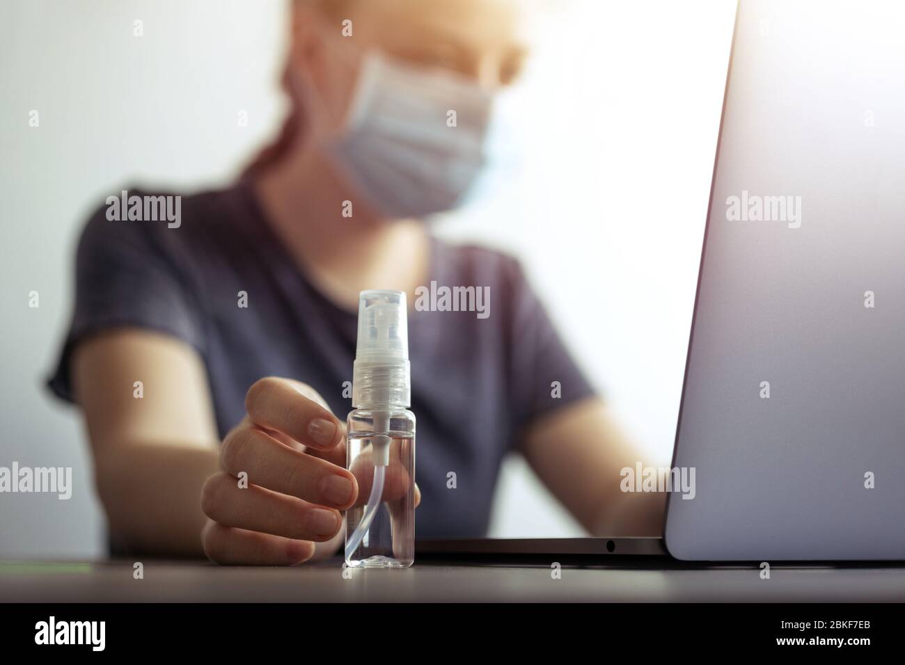 Femme au masque et à la main avec un flacon d'alcool désinfectant près d'un ordinateur portable. Prévention du coronavirus covid-19 en milieu de travail ou à domicile pendant la quarantaine Banque D'Images