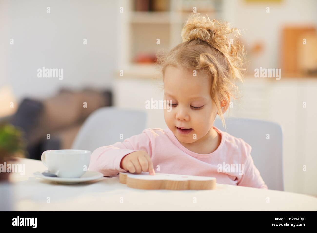 Portrait horizontal moyen gros plan de petite fille caucasienne avec des cheveux onagés blond portant un sweat-shirt rose pâle assis à la table le matin Banque D'Images