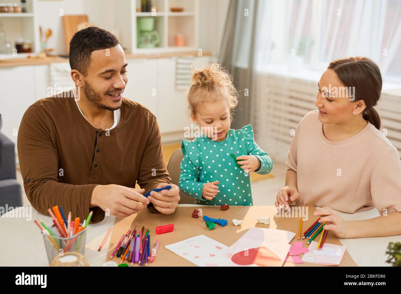 Les jeunes parents adultes passent avec leur petite fille mignonne assise ensemble à des modèles de fabrication de table utilisant de la pâte de jeu colorée Banque D'Images