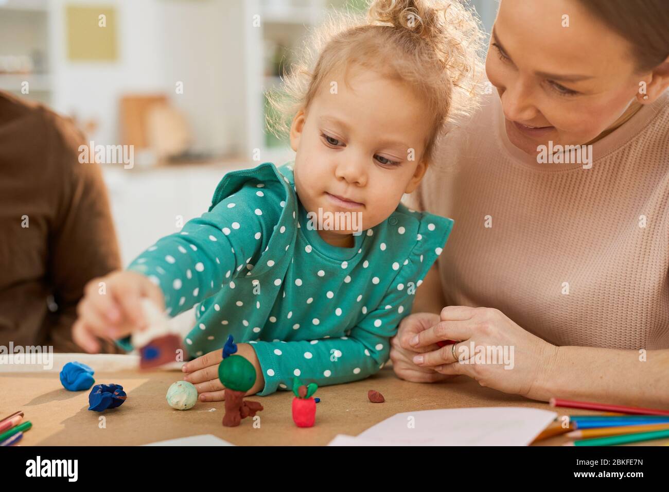 Portrait moyen horizontal de petite fille portant des pois turquoise ensemble assis à table avec les parents à faire des formes à l'aide de pâte de jeu colorée Banque D'Images