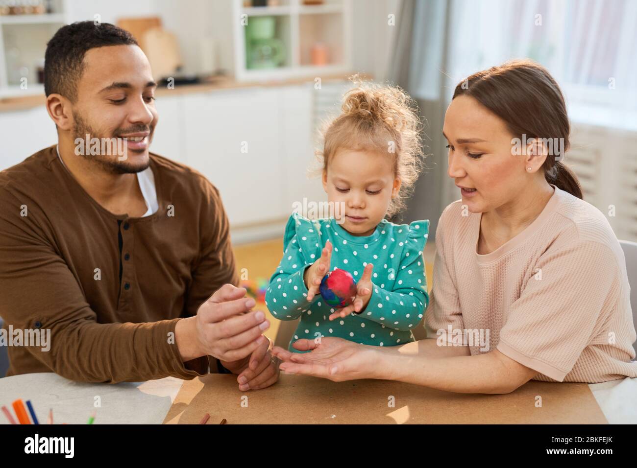 Les jeunes parents assis à la table aident leur petite fille à faire de la boule d'argile de modélisation colorée, tir horizontal Banque D'Images