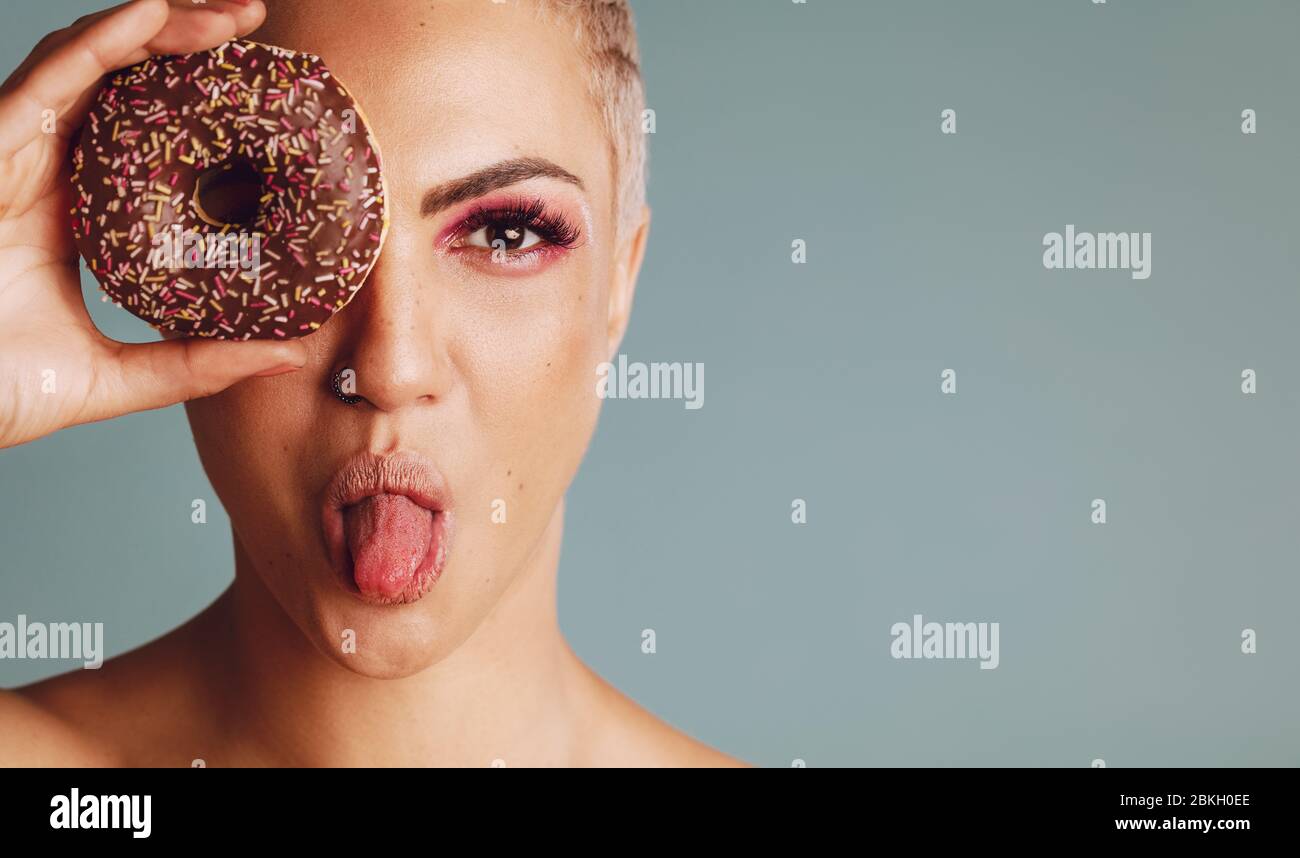 Gros plan d'une femme avec des cheveux courts regardant à travers un beignet et de sortir de sa langue. Belle femme avec un beignet sur fond gris. Banque D'Images