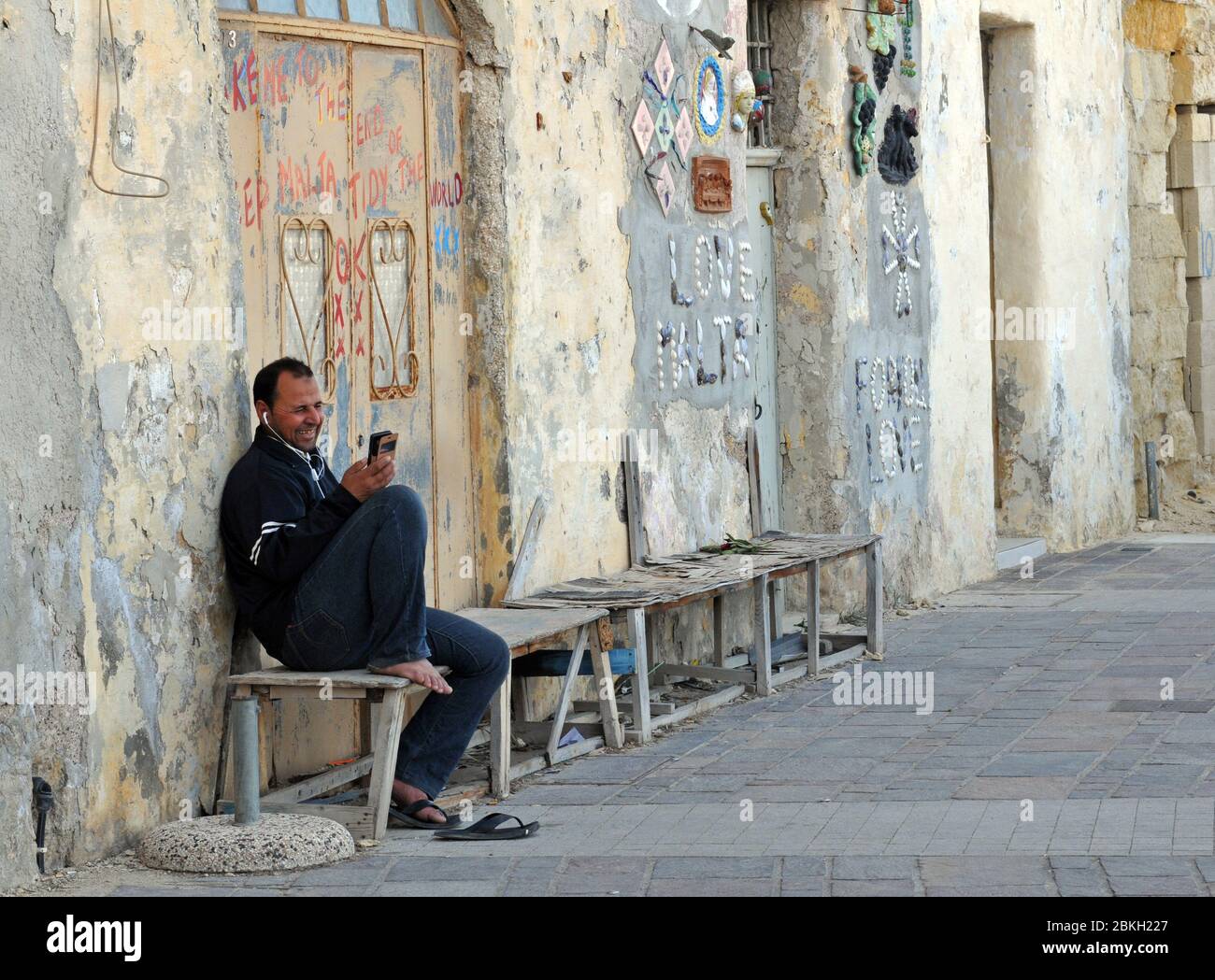 Un homme sourit alors qu'il parle sur un téléphone portable alors qu'il est assis sur un banc dans le village de Marsaxlokk, à Malte. Des œuvres d'art en mosaïque décorent les murs voisins. Banque D'Images