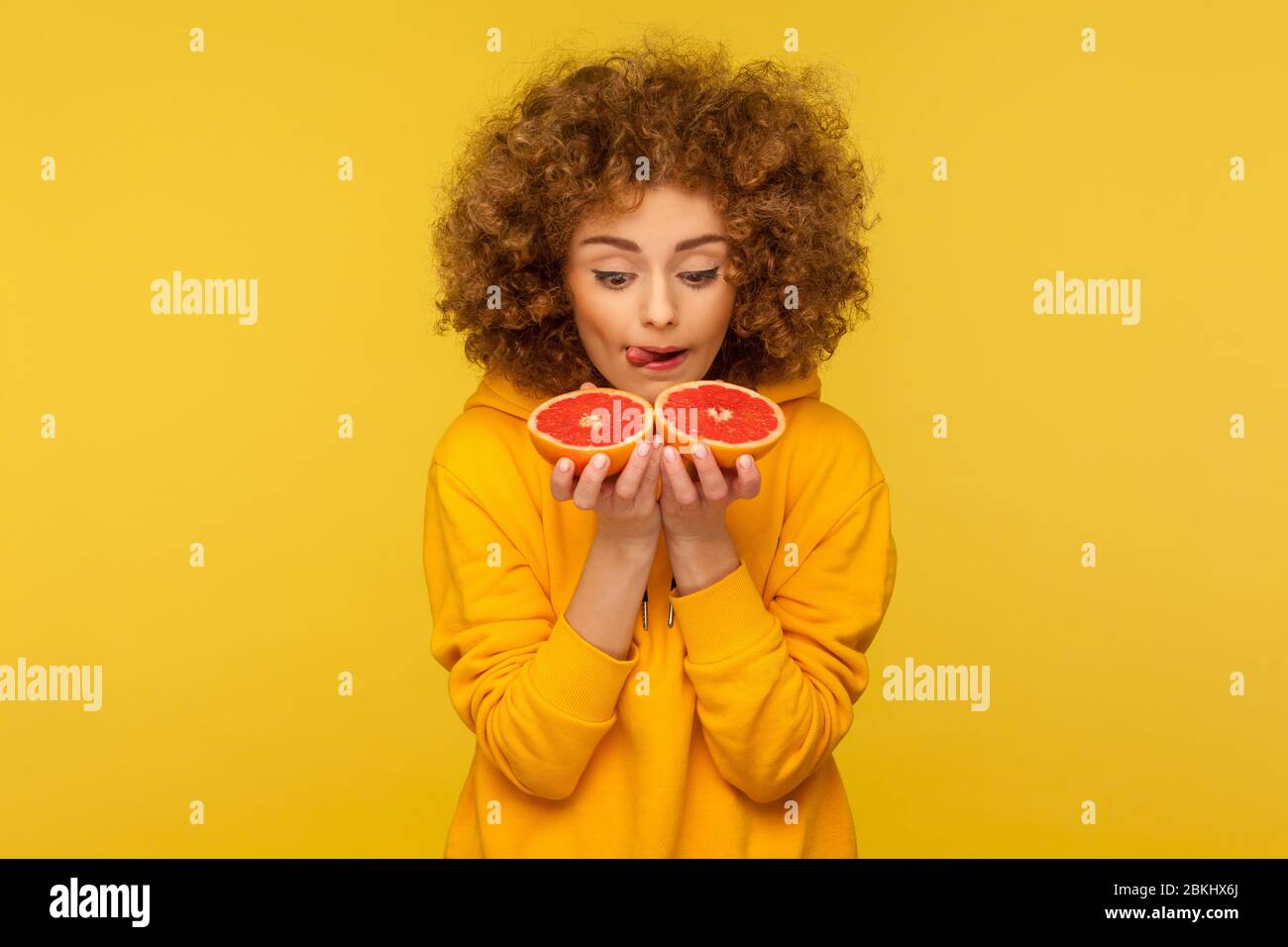 Femme gaie lumineuse avec des cheveux bouclés doux regardant la moitié de tranche de pamplemousse avec le désir, rêvant de manger des fruits frais de vitamine savoureux, une alimentation saine. Dans Banque D'Images